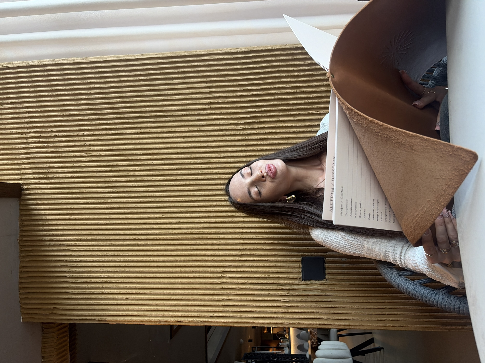 A young woman with long dark hair is seated in a cafe, looking at a menu. She has her eyes closed and her lips puckered, as if she is making a kissy face. She is wearing a white knitted sweater and large gold hoop earrings. The cafe has a modern, minimalist design with a textured yellow wall and white curtains. The woman is holding a menu with a brown leather cover, and some text in Cyrillic is visible on the menu. The overall setting suggests a relaxed atmosphere in a cafe.