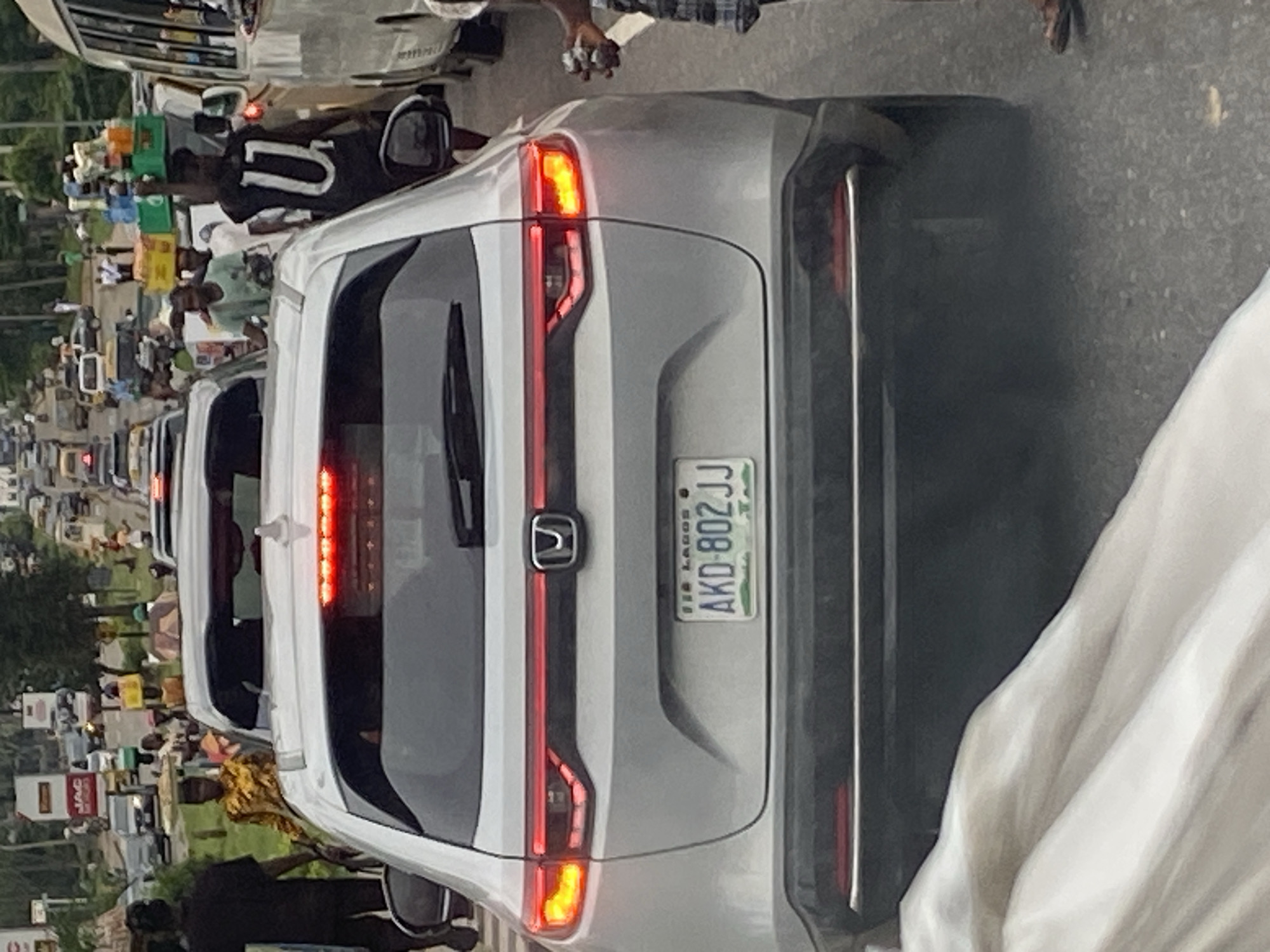 The image shows a traffic jam on a street in Ikeja, Nigeria. The focus is on the rear of a silver Honda SUV. The SUV has its brake lights illuminated, indicating it is stopped. Its license plate reads "AKD-802JJ" and "FED LAGOS".
In the surrounding traffic, a man wearing a black t-shirt with a white "U" logo is visible, carrying several boxes of bottled beverages on his head. Other people are also present, some appearing to be vendors or pedestrians navigating the traffic. Vehicles of various types are seen in the background, contributing to the congestion. The environment appears to be an urban setting with some vegetation visible along the roadside.