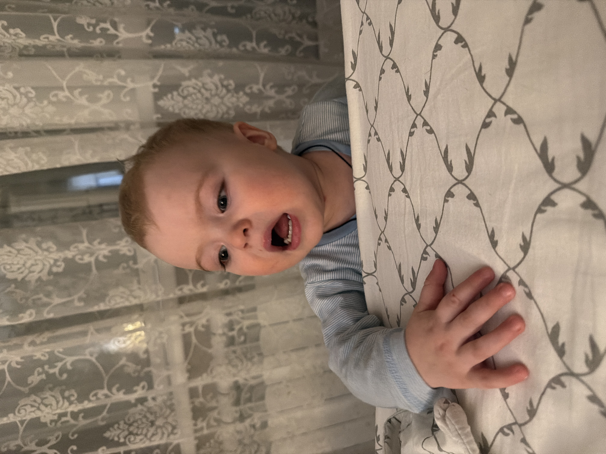 A baby with light hair is looking out from behind a bed in a room. The baby is wearing a long-sleeved, blue and white striped shirt. The baby's mouth is open. The bed is covered with a patterned white and gray sheet. In the background, there is a patterned white curtain. The location is unknown.