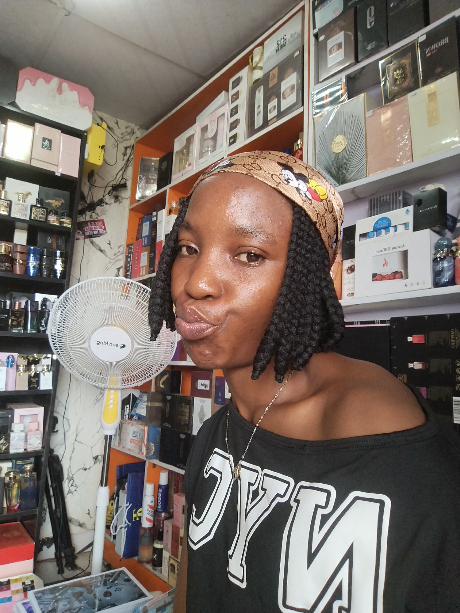 A young Black woman with dark skin and long, braided hair, styled with a brown and tan headscarf featuring a Mickey Mouse design, is captured in a medium close-up shot. She is facing the camera, her lips pursed. She is wearing a black t-shirt with a white "NYC" graphic design and a thin silver chain.
The background suggests she is in a retail environment, possibly a perfume shop. Shelves filled with various boxes and bottles are visible on the left and right sides of the frame. On the left, there are shelves stocked with perfume bottles of different shapes and colors, along with some boxed perfume sets. A tall, white and yellow oscillating fan is positioned behind her, to her left. To the right of the fan, there are more shelves displaying perfume boxes.
The environment appears to be indoors, with a white ceiling and walls. The lighting is bright, illuminating the scene. The overall impression is that of a retail store, possibly in Ugbowo, Nigeria, given the prompt.