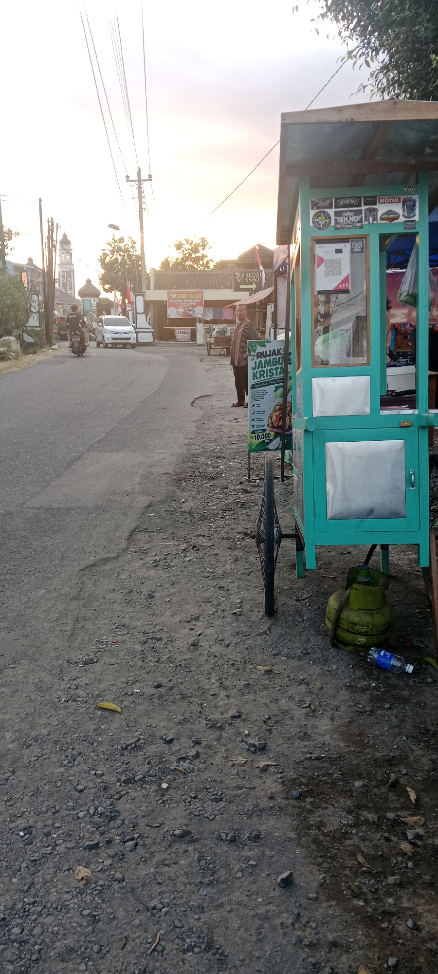 The image depicts a street scene in Maguwoharjo, Indonesia, with a food cart positioned on the right side of the frame. The food cart is turquoise and has a display area with various items, including what appears to be food and drinks. Stickers with logos are affixed to the cart's exterior. A QR code is visible on the cart's front panel. A gas cylinder is placed on the ground near the cart, along with a plastic water bottle. A bicycle wheel is attached to the cart.
In the background, a road curves to the left. A white car is traveling on the road, and a motorcycle with a rider is also present. Several buildings line the street, including one with a prominent tower that resembles a minaret. Signs with text, such as "KEDAI SABO" and "AYAM SEGAR KARUNIA," are displayed on the buildings. A man in a striped shirt is standing near the food cart. The sky is overcast with the sun low on the horizon, suggesting either sunrise or sunset. The ground is covered with gravel and some scattered leaves. Electrical wires are strung across the sky, supported by utility poles. The overall environment appears to be an outdoor, semi-urban setting.