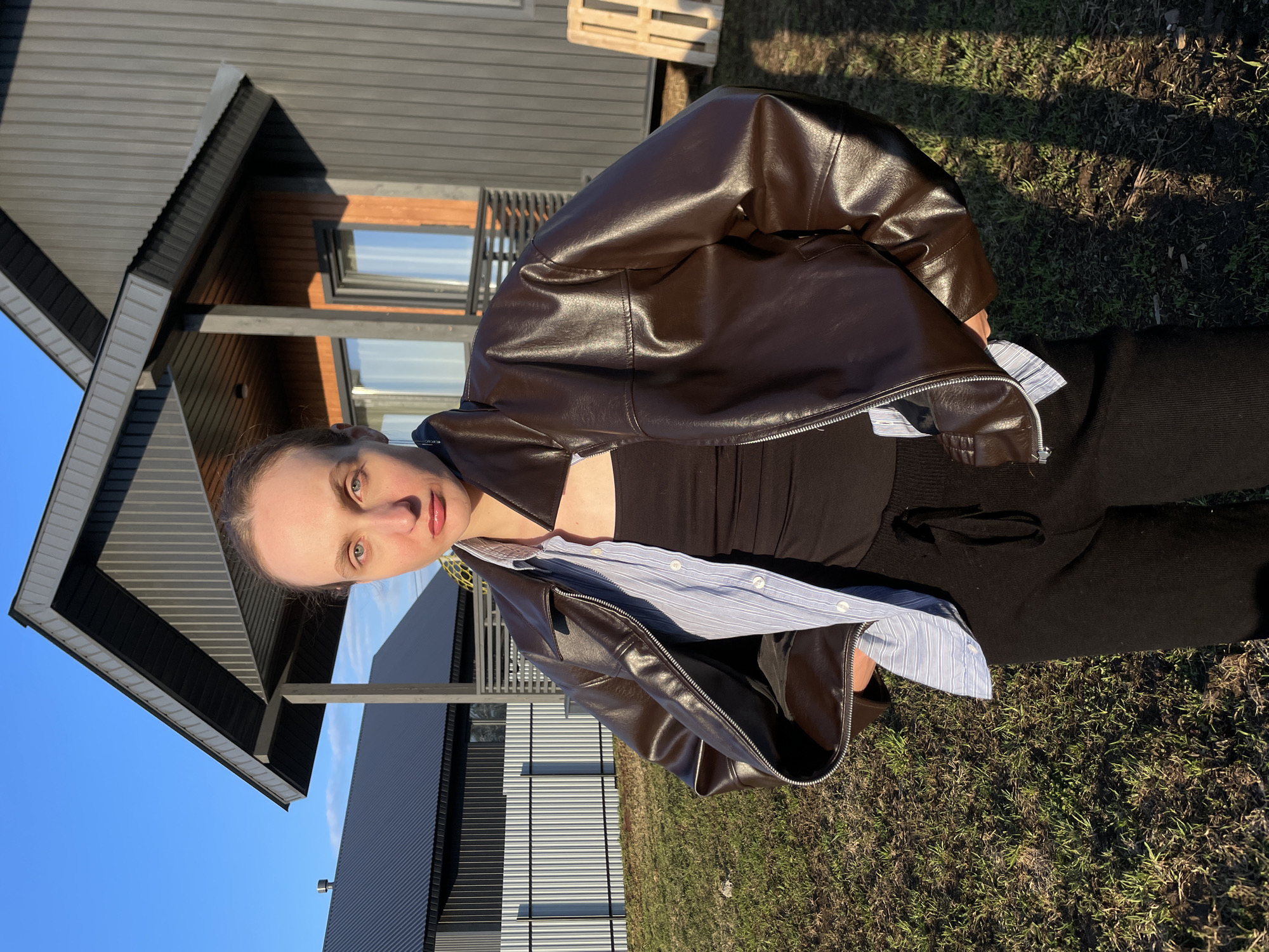 A woman is captured in a close-up, chest-up shot outdoors, looking directly at the viewer with a neutral expression. She wears a dark brown leather jacket, which is partially draped over her shoulders, revealing a black top and a light blue and white striped long-sleeve shirt underneath. Her black pants are visible from the waist down.
The background features a modern-style house with dark grey corrugated metal siding and sections of light-colored wood paneling, particularly around the windows. The architecture includes angular rooflines and structural beams. The sky is clear and bright blue. The ground is covered in a mixture of green and dry grass, displaying long shadows that suggest the photo was taken during early morning or late afternoon sunlight. A wooden pallet is visible in the upper right background. There is no visible text in the image. The setting appears to be a residential or semi-rural area, consistent with Podshivalovo, Russia.
