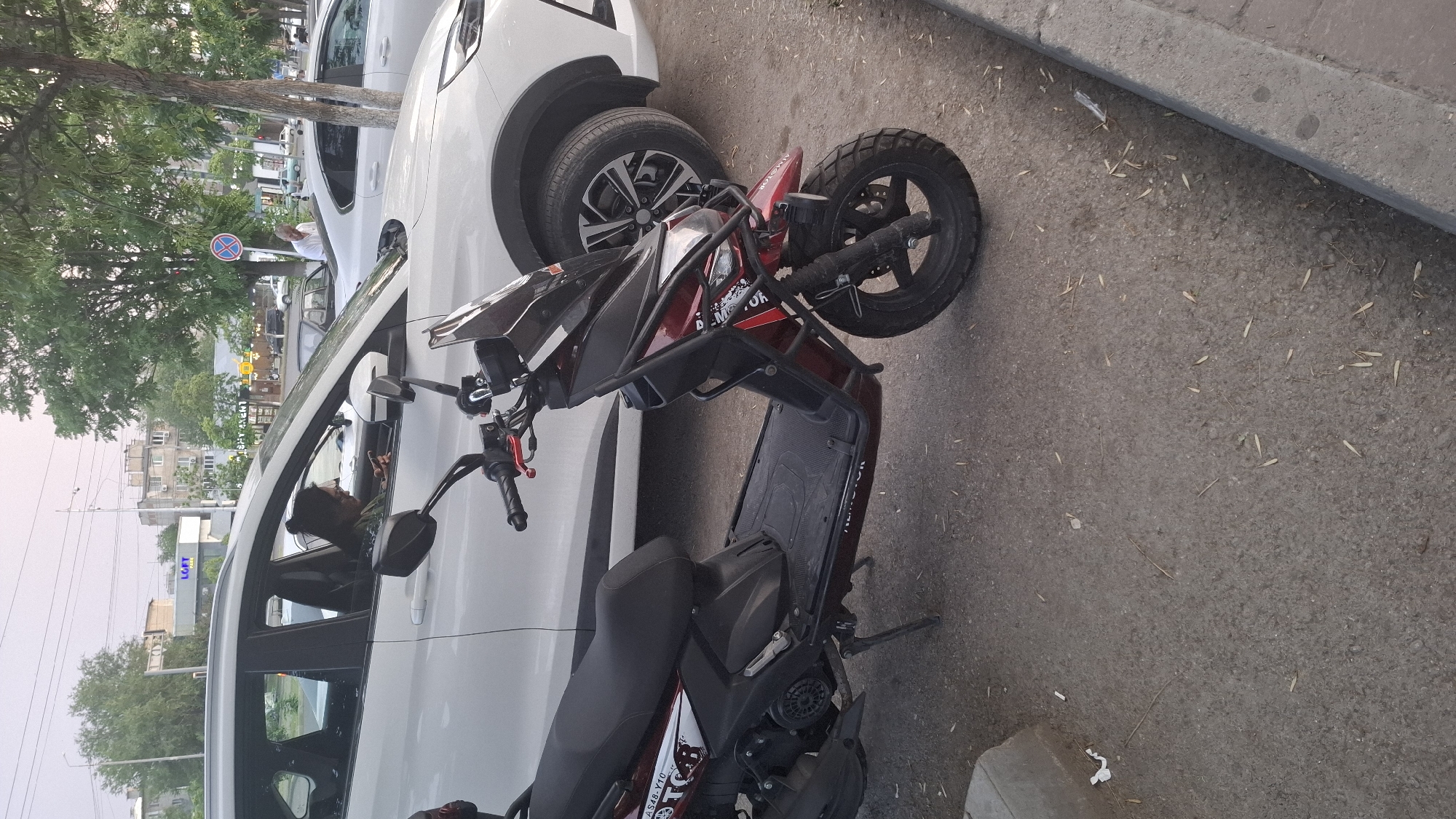 A dark red and black motor scooter is parked next to a white SUV on an asphalt street in Shymkent, Kazakhstan. The scooter is positioned at an angle, with its front wheel turned slightly to the left. It has a prominent black metal frame around the handlebars and front of the vehicle. The seat is black, and there are some white decals with red lettering on the body of the scooter. The SUV is parked to the left of the scooter, partially obscuring it. The SUV is white and appears to be a modern model. Its front wheel is visible, showing a silver rim with black accents.
In the background, behind the SUV, there is a tree with green foliage. Further back, buildings and more trees can be seen. A blue and red traffic sign with a white cross symbol is visible on a pole to the left of the SUV. The sky appears overcast, suggesting it might be late afternoon or early evening, or a cloudy day. The lighting is somewhat dim. There is a curb visible in the lower right corner of the image. The ground is paved with asphalt and is scattered with small leaves or debris. Some people are visible through the windows of the SUV. One person in the driver's seat appears to be looking towards the side. Another person is visible in the passenger seat of the SUV, looking out the window. Text on the scooter is partially visible, including "DEMO-MOTO" and what looks like "XTO".