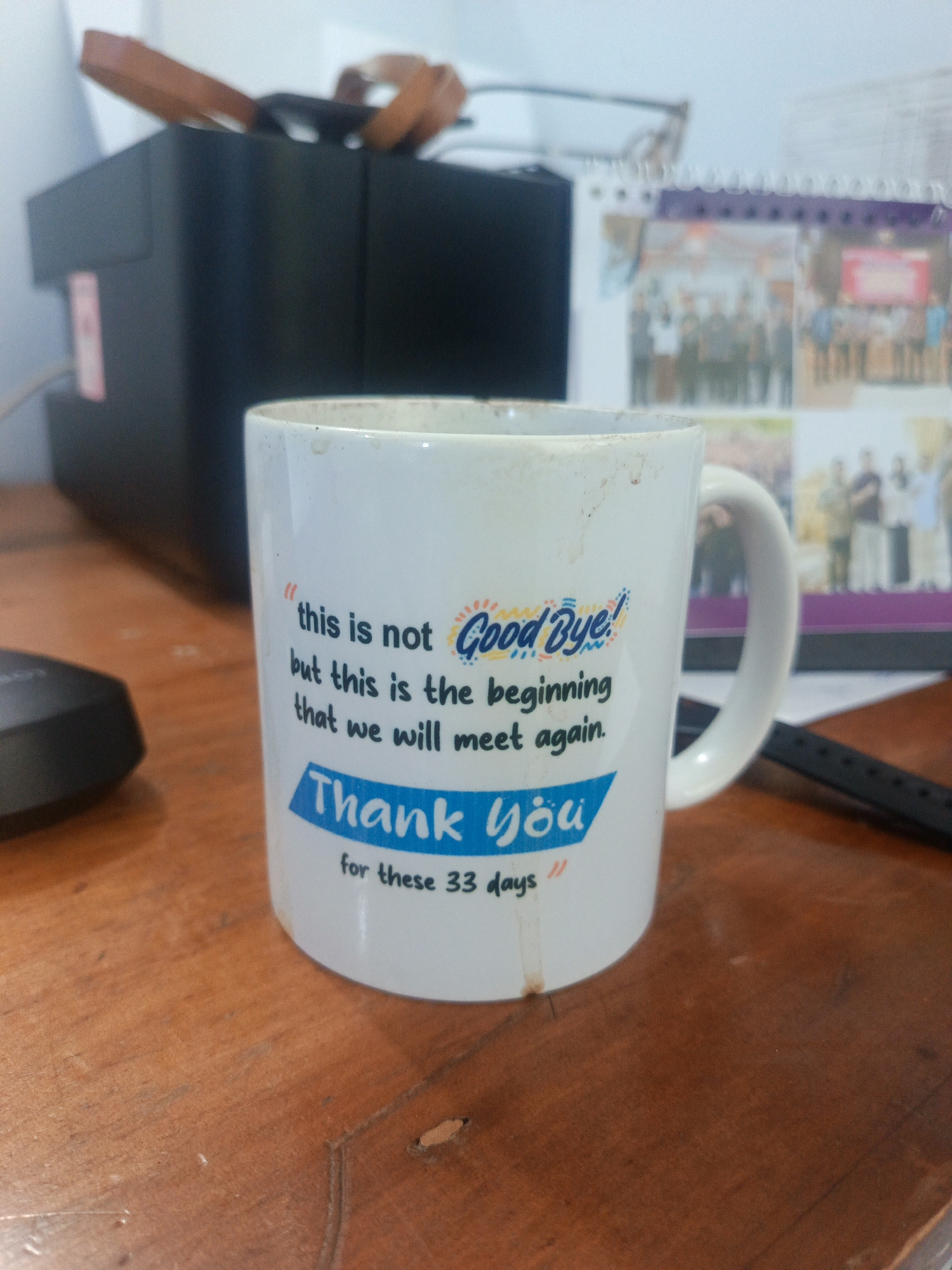 An indoor, medium shot captures a white ceramic mug on a wooden surface. The mug, positioned slightly off-center to the right, features blue and orange text. The text reads: "this is not Good Bye! but this is the beginning that we will meet again. Thank You for these 33 days". A dark object, possibly a mouse, is visible to the left of the mug. In the background, a black office machine, a calendar with photos of people, and a pair of glasses are out of focus. The wooden surface appears worn with visible grain and subtle indentations. There are no people visible in the image. The lighting suggests an indoor environment, possibly an office or a home desk. The overall impression is that of a personal item left on a desk. No specific information about the city of Gorontalo, Indonesia, can be inferred from this image.