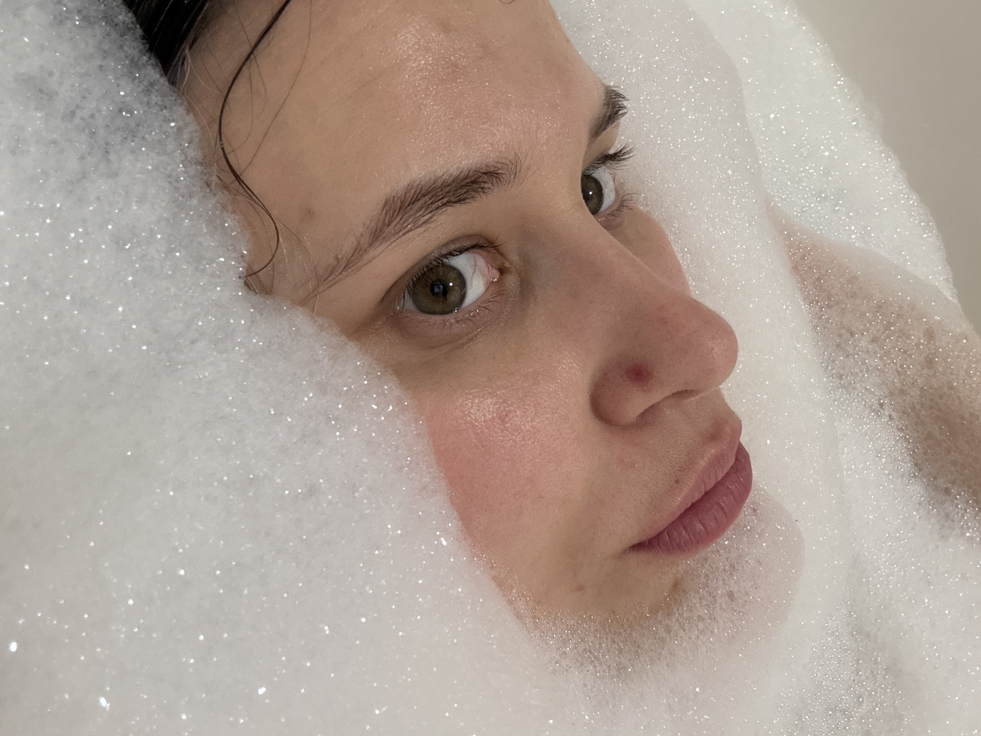 A woman is partially submerged in a bathtub filled with dense white foam. Only her face and a small part of her chest are visible above the foam. Her face is turned slightly to her right. She has fair skin with a slightly flushed cheek and a small red blemish on her nose. Her eyes are brown and she has dark eyebrows and eyelashes. Dark hair is visible near her forehead, partially covered by the foam. The foam is evenly distributed around her face, obscuring the contours of her body. The scene appears to be indoors, likely a private bathroom. There is no information about the location of Chaikovsky, Russia, from this image.