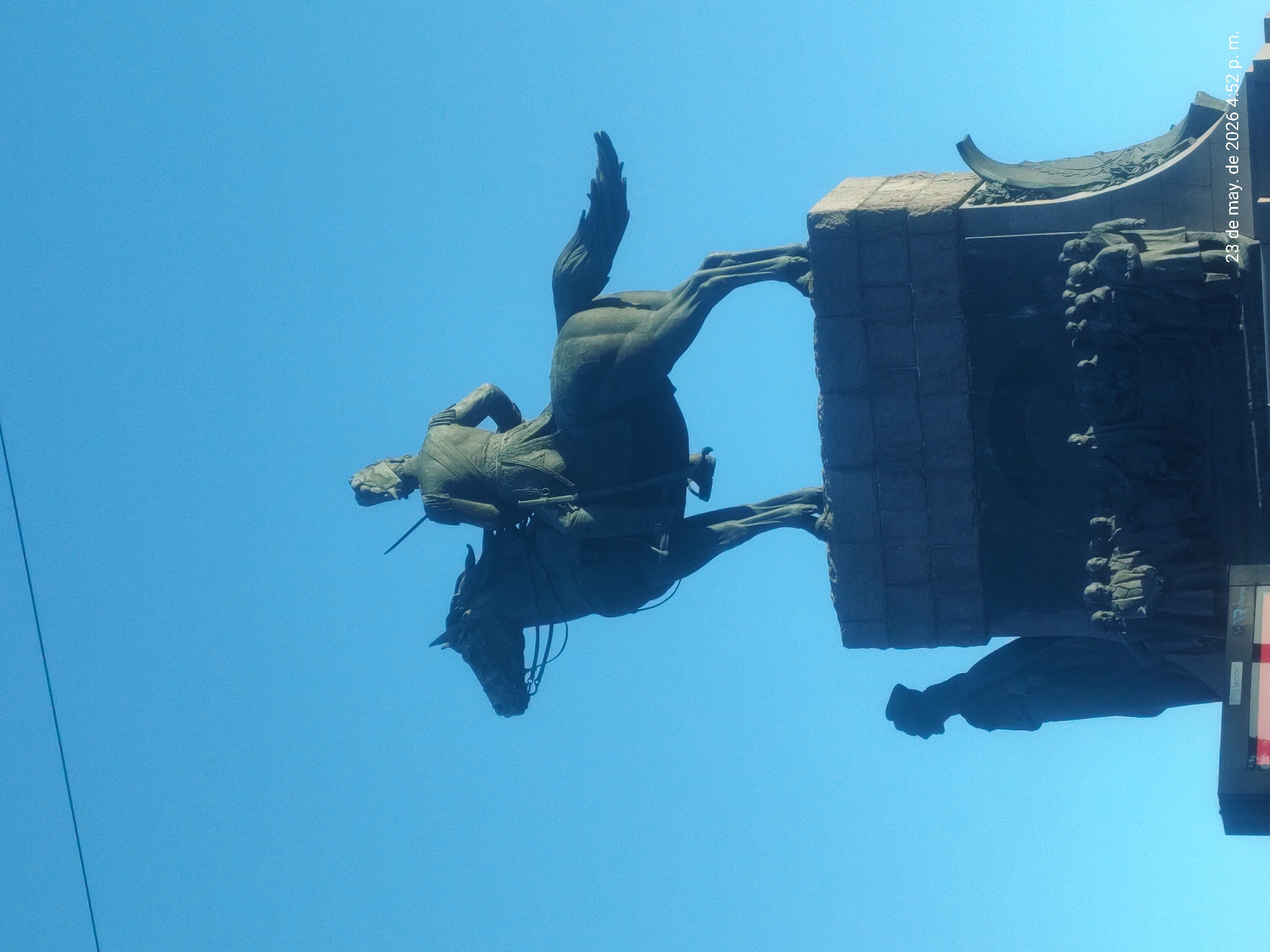 The image captures an upward view of the equestrian monument of General José de San Martín, located in Plaza San Martín, Buenos Aires, Argentina. The bronze statue depicts San Martín on horseback, the horse rearing with its front legs perched on the edge of the monument's stone pedestal. The pedestal is adorned with relief sculptures. Above, the sky is clear and blue. A date, “23 de may. de 2026 4:52 p. m.” is visible vertically along the right edge of the image.