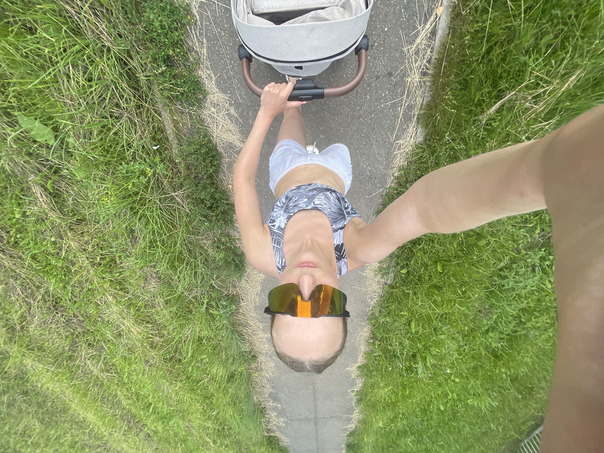 In a high-angle shot looking down, a person is seen walking on a paved path, pushing a light-colored baby stroller. The person is wearing a grey and white patterned sports bra, white shorts, and large, reflective orange sunglasses. They are holding the stroller's handle with their right hand, and their left arm is extended towards the camera, implying they are taking a selfie. The path is bordered by lush green grass, with some dry, straw-like grass also visible. The scene appears to be outdoors, in an area that could be a park or a residential street, potentially within the village of Podshivalovo, Russia, given the context. The lighting suggests it is daytime.