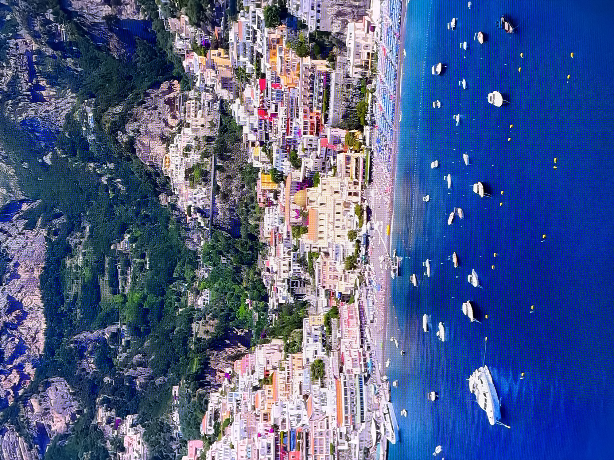 This is an aerial view of a coastal town nestled against a steep, rocky mountain. The town is characterized by densely packed buildings with colorful facades in shades of pink, yellow, white, and orange, many with red-tiled roofs. These structures cascade down the mountainside towards a vibrant blue body of water. A beach lined with rows of blue and white lounge chairs is visible at the base of the town. Numerous boats, ranging from small dinghies to larger yachts, are scattered across the water, some moored and others appearing to be underway. The water itself is a deep, rich blue, reflecting the bright sunlight. The mountainous terrain above the town is covered with lush green vegetation and rugged rock formations. There are no people clearly visible in the image. The scene depicts a popular tourist destination with a characteristic Mediterranean aesthetic. The city of Chirchik, Uzbekistan is not depicted in this image.