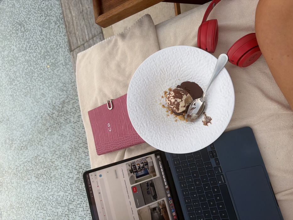 This image depicts a poolside lounge area in Dubai, United Arab Emirates. A dark blue laptop is open on a light-colored, possibly beige, cushioned lounge chair. The laptop screen displays a Pinterest interface with various images, including a grey vehicle and interior design concepts. To the right of the laptop, a pink, crocodile-embossed notebook or journal featuring the silver initials "C.D." and a silver clasp is placed on the cushion. A white, textured ceramic bowl is centrally positioned, holding a dessert consisting of a scoop of marbled ice cream on a bed of chocolate sauce, surrounded by biscuit crumbs. A silver spoon rests within the bowl, partially immersed in the chocolate. A pair of red Beats by Dre wireless headphones lie on the cushion to the right of the bowl. The lower right corner reveals a tanned leg belonging to the person occupying the lounge chair. In the background, a light blue mosaic-tiled swimming pool is visible, along with a grey stone border marking the pool's edge. A partial view of a wooden tray or table is visible on the far right.