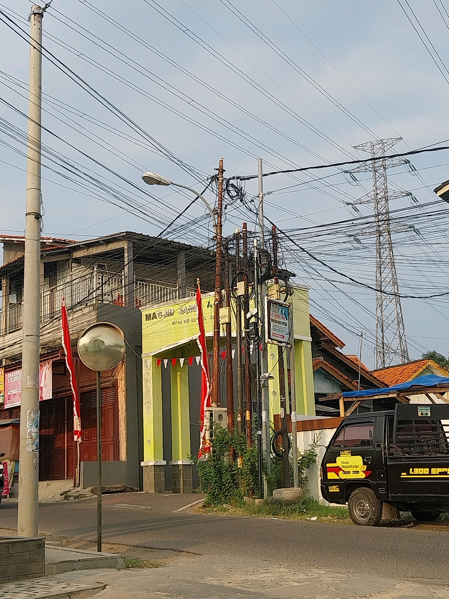 The image captures a street scene in Cirebon, Indonesia, under a hazy sky. A concrete utility pole stands prominently on the left, draped with a dense network of power lines. To its right, a building with a lime green facade and dark gray columns is visible, adorned with red and white triangular banners. Above the building, a second story with a balcony and metal railings is partially obscured by more wires. A large, convex mirror is mounted on a pole in front of the building, reflecting a distorted view of the surroundings. Further to the right, a black pickup truck is parked on the asphalt road. Behind the truck, a tall electrical tower is silhouetted against the sky, also surrounded by a complex web of wires. Red flags hang from the building on the left. The overall impression is one of urban infrastructure and everyday life in a densely wired environment.