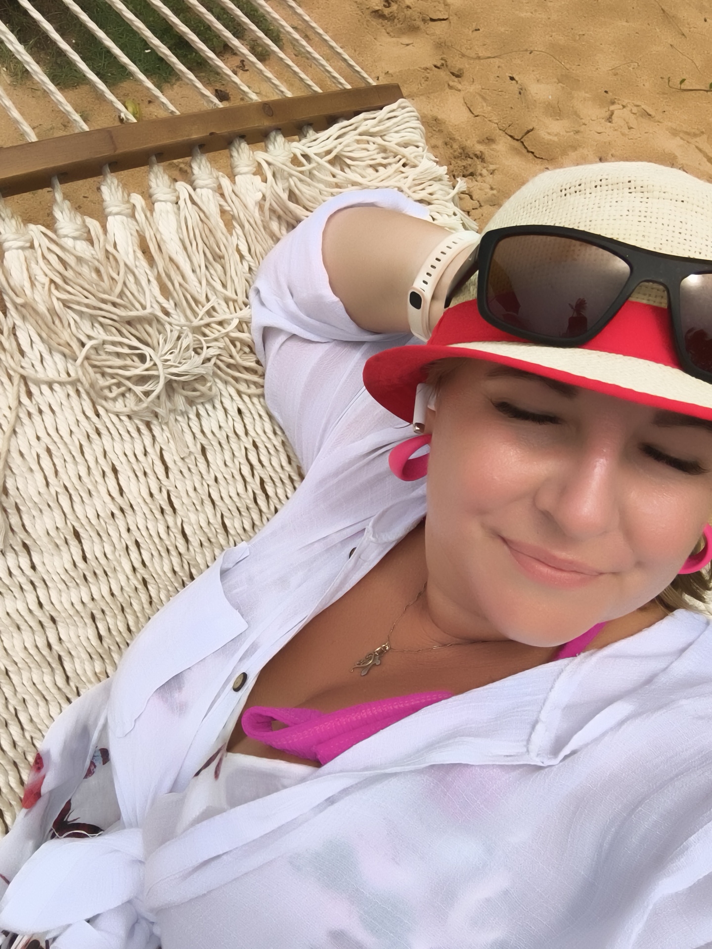 A woman lies in a hammock on a sandy beach in Phú Quốc, Vietnam, taking a selfie. She is wearing a white sheer shirt with a floral pattern, a bright pink bikini top, a red and white hat, and sunglasses. She also has on large pink hoop earrings and a delicate gold necklace. The hammock is made of thick white rope. In the background, there is sand and some greenery.