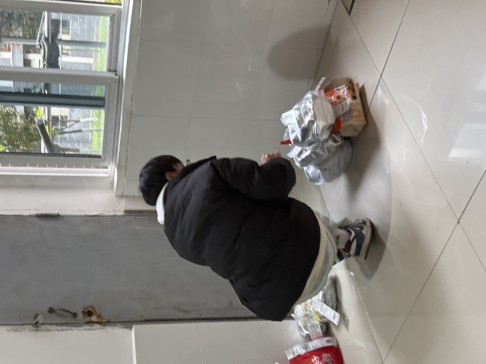 An individual is depicted crouching on a light-colored tiled floor within an interior space in Jiangjin, China. The person, viewed from behind, wears a black puffer jacket with a white collar, light-colored pants, and athletic shoes with dark blue, white, and red accents. They are oriented towards the right of the frame, bending over a collection of packaged goods.
The items on the floor include several packages wrapped in silver-colored plastic or foil, some appearing as individual units, along with a rectangular cardboard box featuring red and orange branding, possibly depicting a cartoon tiger. A small red clip or similar object is visible among these items. A red bag with white Chinese characters is partially visible on the far left. The individual's hands are positioned near the center of the packaged goods, suggesting an action of organizing or retrieving items.
The immediate environment features plain white walls. To the left, a light-grey metal door with a simple handle is visible, exhibiting some rust or discoloration near the frame and a small green object affixed near the top. To the right, a multi-pane window with white frames provides a view of an outdoor area. Through the window, green foliage, a paved or grassy area, a grey brick-patterned building, and a fence structure are discernible, indicating an urban or suburban setting. Shadows are cast on the tiled floor, suggesting an overhead or side light source.