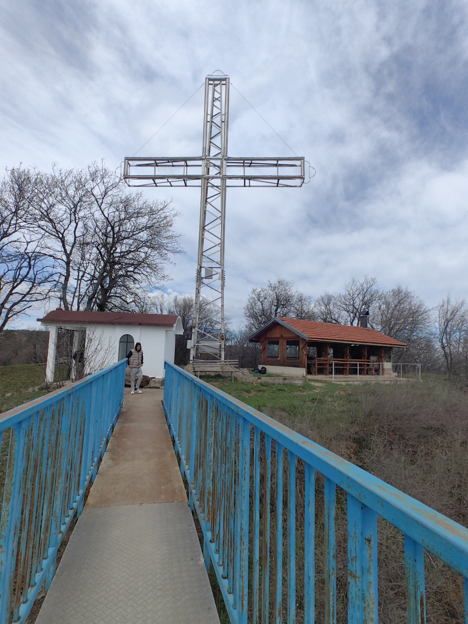 The image depicts a landscape in Vrabevo, Bulgaria, dominated by a large metal cross structure. A blue metal bridge leads towards a small white building with a red roof. A person is standing on the bridge, near the white building. A wooden building with a red tile roof is positioned to the right, further in the background. The surrounding environment consists of bare trees, bushes, and grass. The sky is overcast with patchy clouds.