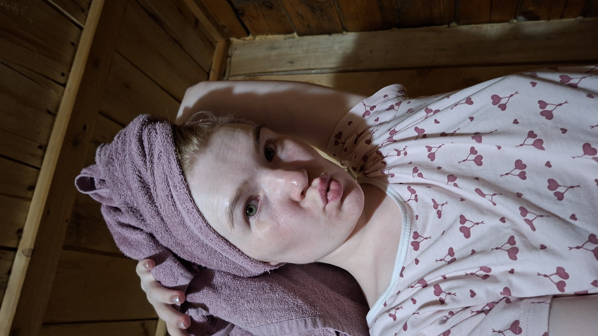 The image shows a pale-skinned person with a towel wrapped around their head, lying in what appears to be a wooden sauna or cabin. The person is wearing a pink and white patterned pajama top. The background is comprised of wooden planks, indicative of the interior structure. The person is looking at the camera with their lips pursed. There is no clear indication of the location being Большой Букор, Russia, from the provided image.