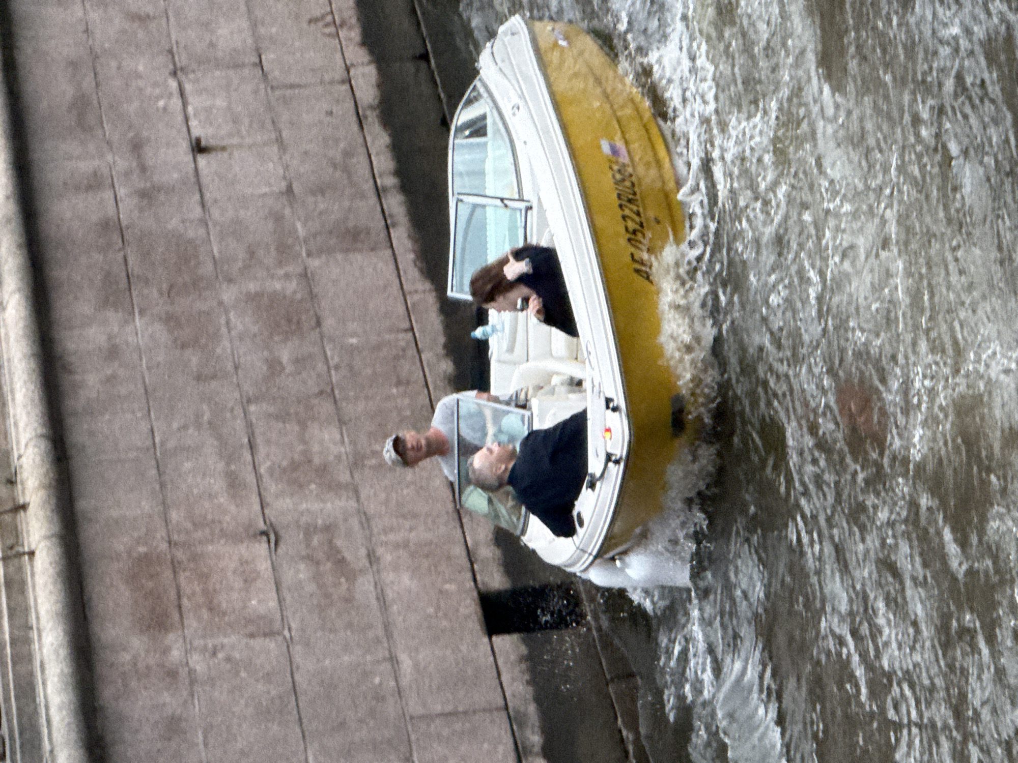 A yellow and white motorboat is navigating through choppy brown water, likely a canal or river in Saint Petersburg, Russia, given the presence of a brick embankment in the background. The boat is occupied by three individuals. A man in a grey t-shirt and a baseball cap is operating the boat from the rear. In the front passenger seats are two individuals. A man in a dark suit is seated on the left, facing away from the camera. A woman with long brown hair, wearing a dark top, is seated on the right, also facing away from the camera. The boat has the license plate "AE 0522 RUS61" visible on its side. The water is turbulent, creating spray around the boat, indicating it is moving at a moderate speed. The embankment is constructed from large, rectangular grey stones.
