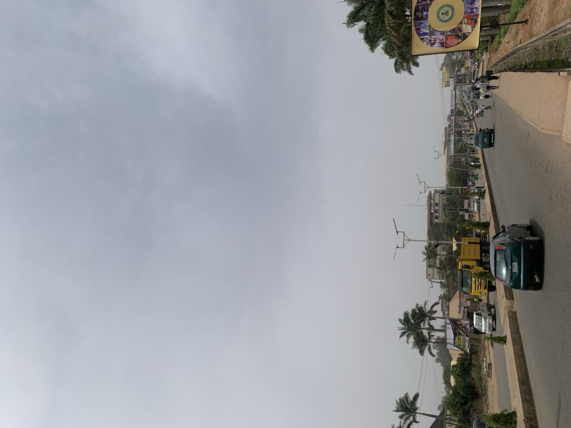 The image presents a daytime street scene in Jos, Nigeria, observed from a slightly elevated perspective overlooking a paved road extending into the distance. The sky is uniformly gray and overcast or hazy, indicating diffused light without direct sunlight.
The main subjects include the road, which appears moderately busy with vehicular traffic, and its adjacent sidewalks. In the foreground on the right, a dark green car, possibly a hatchback, is visible moving or parked, with a yellow dump truck behind it. Further along the road, various other vehicles, including cars and smaller commercial vehicles or motorcycles, are present. Pedestrians can also be seen walking on the concrete sidewalk on both sides of the street.
Numerous tall palm trees are a dominant feature, lining the road on both sides and contributing to the urban landscape. Beyond the trees and traffic, several low-to-medium-rise buildings are visible in the background, partially obscured by the atmospheric haze. Modern street light poles with multiple arms are installed along the road.
A notable detail is a large circular banner prominently displayed on a pole on the left side of the road. This banner has a green outer ring and a yellow-gold inner circle. Visible text on the banner reads, "PLATEAU STATE OF NIGERIA" and "GOVERNMENT OF PLATEAU STATE," surrounding a collage of smaller images. Further down the street, another green and white banner or billboard is partially legible, showing phrases like "SPECIAL ANNOUNCEMENT" and "SCHOOL RESUMPTION." The overall scene depicts typical daily activity on a city thoroughfare.