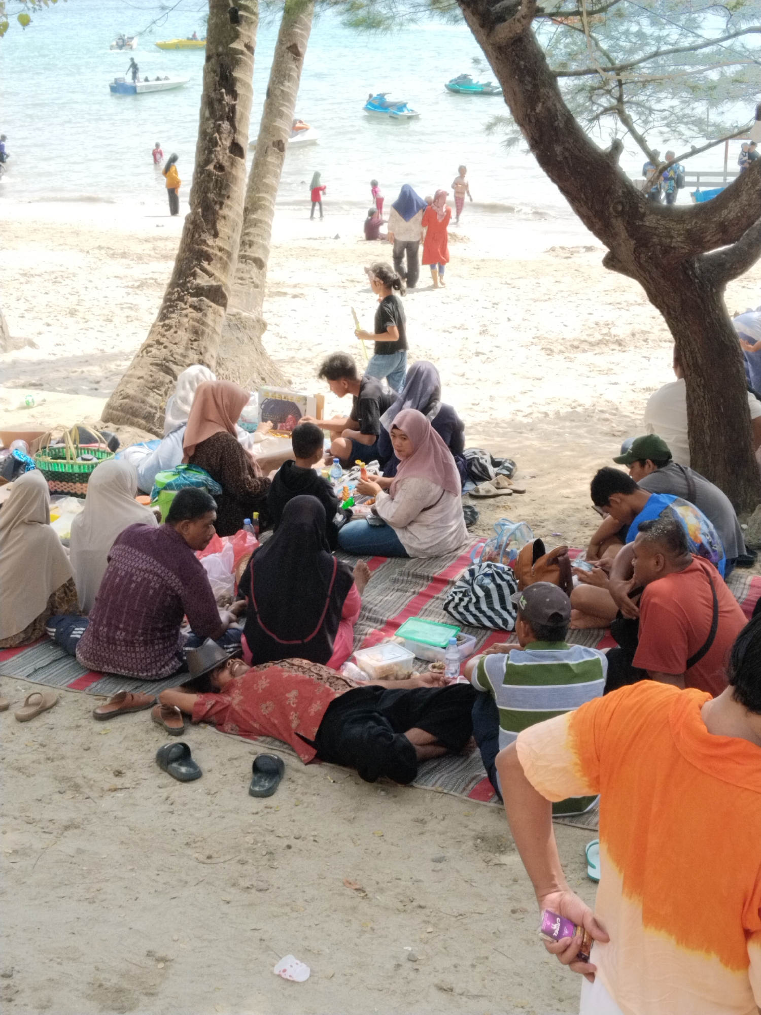 The image depicts a crowded beach scene in Prigi, Indonesia. In the foreground, several people are seated or lying on mats on the sandy shore, appearing to be picnicking or relaxing. They are dressed in casual clothing, with many women wearing headscarves. Objects scattered around include picnic baskets, food containers, and footwear. In the background, the ocean is visible with several boats and jet skis. People are also seen walking along the shoreline and wading in the water. Palm trees and a larger tree with branches loom over the group, providing shade. The overall atmosphere suggests a lively gathering on a sunny day at the beach.