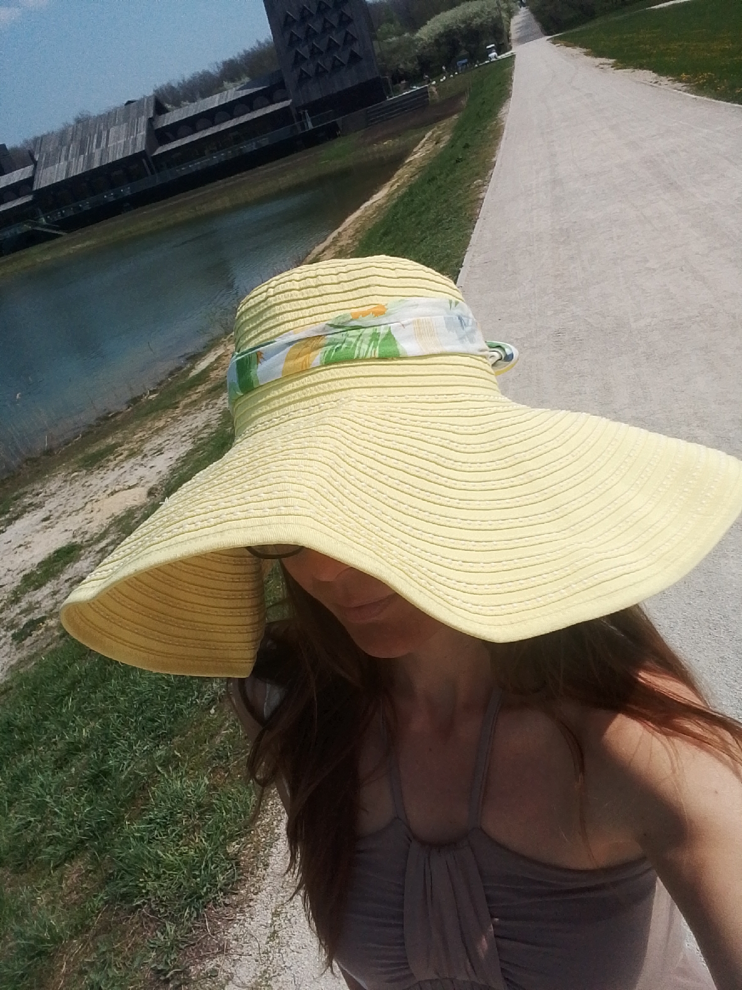 A woman with long brown hair is wearing a wide-brimmed yellow hat with a floral print band. She is standing on a path that extends into the distance. To her left is a body of water, and behind it, a large, modern building complex with a distinctive tower is visible. The location is Каменка, Russia.