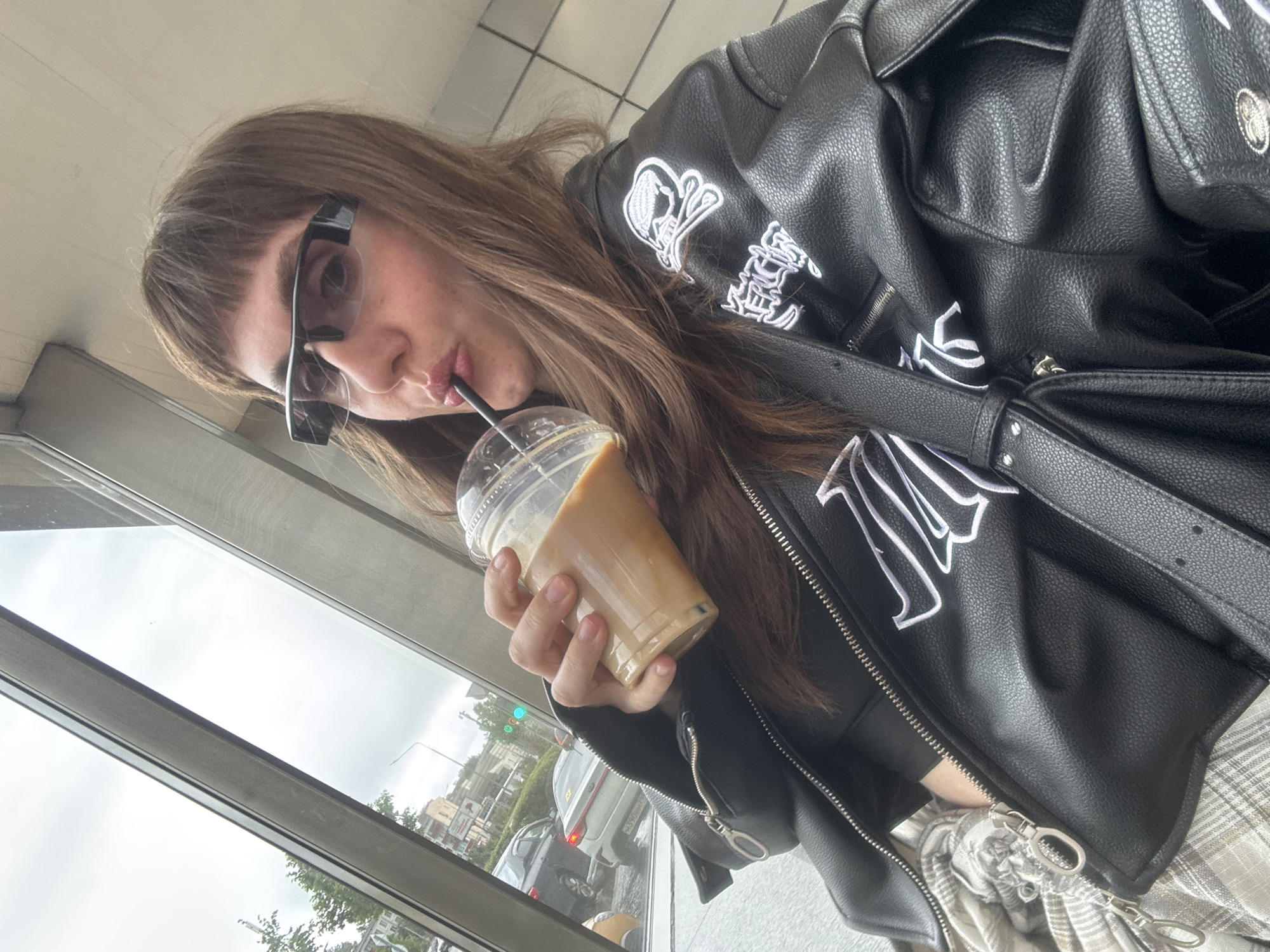 A person is drinking from a clear plastic cup with a straw, likely coffee or a similar beverage. They are wearing sunglasses and a black leather jacket with white graphic designs on the sleeves and chest. A black bag is slung over their shoulder. The background shows a view of a street with vehicles and buildings visible through a window. While the scene is urban, there is no specific information to confirm its location as Moscow, Russia.