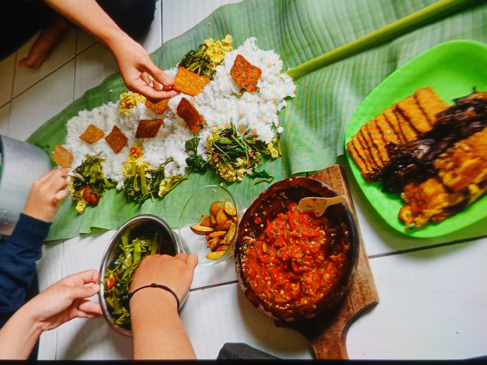 In a domestic setting, likely within the city of Setia Bakti, Indonesia, a group of individuals is sharing a communal meal served on a banana leaf laid out on a tiled floor. Three sets of hands are visible, actively participating in the meal. One hand, adorned with a dark bracelet, is reaching into a metal bowl containing stir-fried green vegetables. Another hand is picking up pieces of fried tempeh from a vibrant green plastic plate. A third hand is taking a piece of fried tempeh from the main serving. The central focus of the meal is a mound of white rice, interspersed with more stir-fried vegetables and what appear to be square pieces of fried tempeh. To the left of the rice, a round wooden mortar filled with a vibrant red chili sambal is prominent, accompanied by a small wooden spoon. A clear glass bowl containing fried potato wedges is situated behind the sambal. In the background, a large metal pot hints at the preparation of the meal. The overall atmosphere suggests a casual, family-style dining experience.