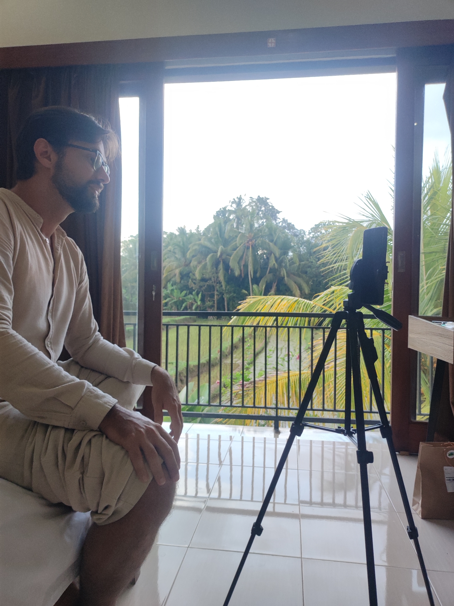 A man with a beard and glasses, dressed in a light-colored, long-sleeved shirt and matching trousers, is seated indoors, looking towards a smartphone mounted on a black tripod. He appears calm and slightly smiling, suggesting he is engaged in a video call, recording, or live streaming.
The setting is a room with large glass doors that open onto a balcony with a black metal railing. Beyond the balcony lies a panoramic view of lush tropical vegetation, including numerous palm trees and what appear to be terraced rice paddies. The bright outdoor light indicates it is daytime, possibly with an overcast sky. The dark brown frames of the doors and windows, along with light-colored curtains, frame the vibrant outdoor scene.
On the tiled floor near the balcony, a brown paper package with a white label containing a barcode and some illegible text is visible. The man's posture is relaxed, with his hands resting on his knee. The combination of the modern indoor space and the verdant, agricultural landscape strongly suggests a tropical location, consistent with Tegallalang, Indonesia.