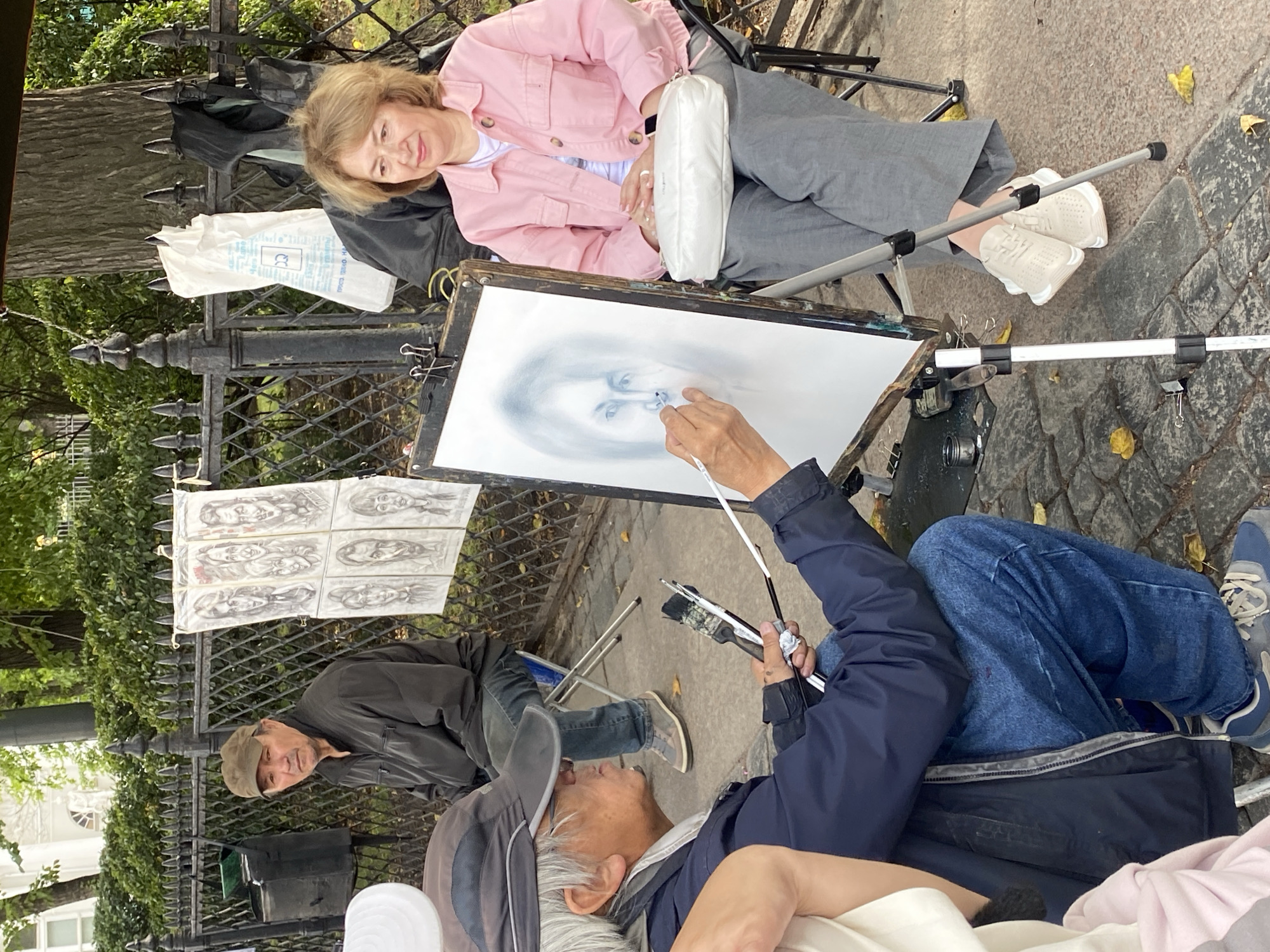 In Saint Petersburg, Russia, an artist is sketching a portrait of a woman sitting on a stool in front of an easel. The artist, an older Asian man wearing a cap and dark jacket, is in the foreground, hunched over the easel, holding brushes. The woman, a fair-skinned woman with blonde hair, wears a pink jacket and gray pants, her hands resting on her lap. Behind them, a black metal fence separates the sidewalk from a grassy area with trees. Several pencil sketches of faces are displayed on the fence. A man in a brown cap and dark jacket sits on a chair near the fence, looking towards the viewer. The ground is paved with cobblestones, with a few fallen leaves scattered around. The overall scene depicts an outdoor art setup in a public space.