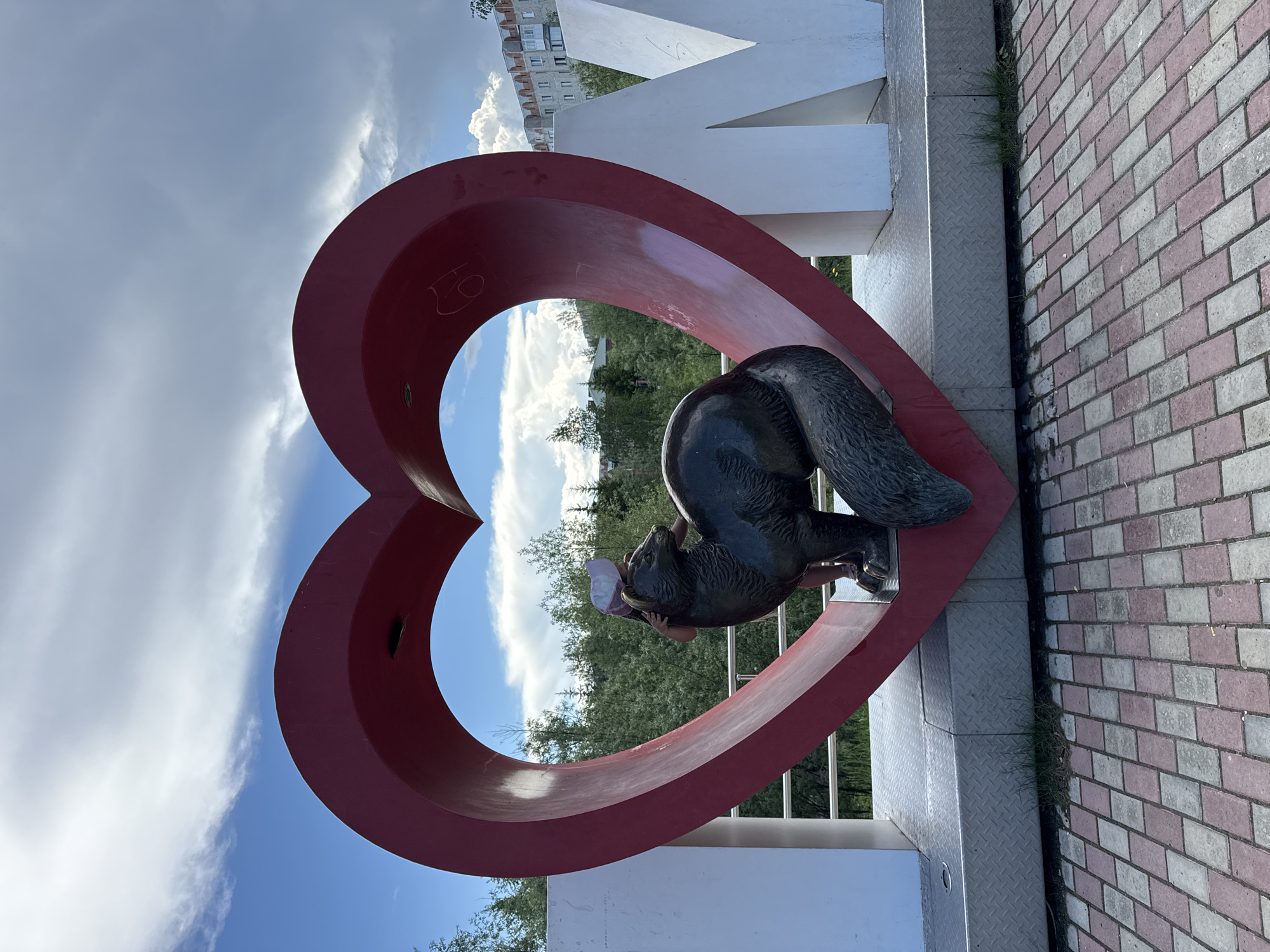 The image shows a large red heart sculpture in the city of Megion, Russia. A statue of a dark-colored animal, possibly a fox or wolverine, is positioned inside the heart. A young person wearing a pink hat is interacting with the animal sculpture, leaning against it with their arms around its neck. In the background, there are trees and a multi-story building. The sculpture is situated on a metal platform, with a patterned brick pathway in the foreground. The sky is partly cloudy with patches of blue.