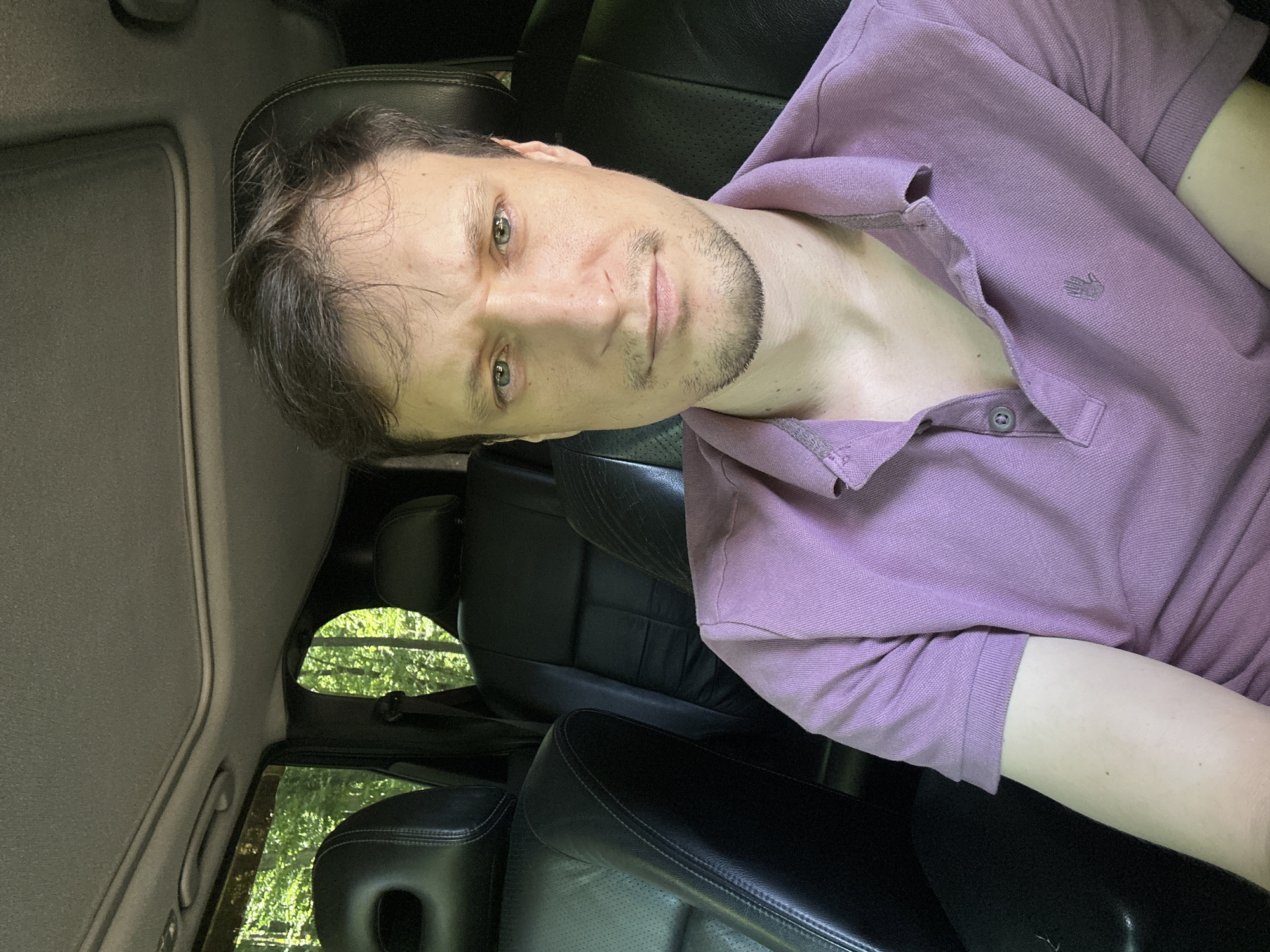 A man is sitting in the driver's seat of a car, looking directly at the camera. The car's interior is visible, with black leather seats and a sunroof. Trees are visible through the car windows, suggesting an outdoor location. The man is wearing a purple polo shirt. The image is a selfie, taken from a slightly elevated angle. The location is not specified in the image or provided context.