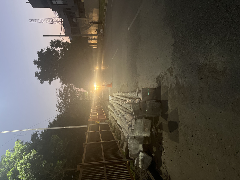 The image shows a street in Sahibzada Ajit Singh Nagar, India. The image is taken at dusk, with the sky a hazy blue. In the foreground, a pile of metal pipes is stacked near a streetlamp. An orange traffic cone is sitting in the middle of the street, which is empty of traffic. The road is paved and appears to be under construction. There is a metal fence along the left side of the road. The background shows trees and buildings in the distance. There is a silhouette of a person standing in the distance. The street is lit by streetlights. The image was taken from the perspective of a person standing on the left side of the road.