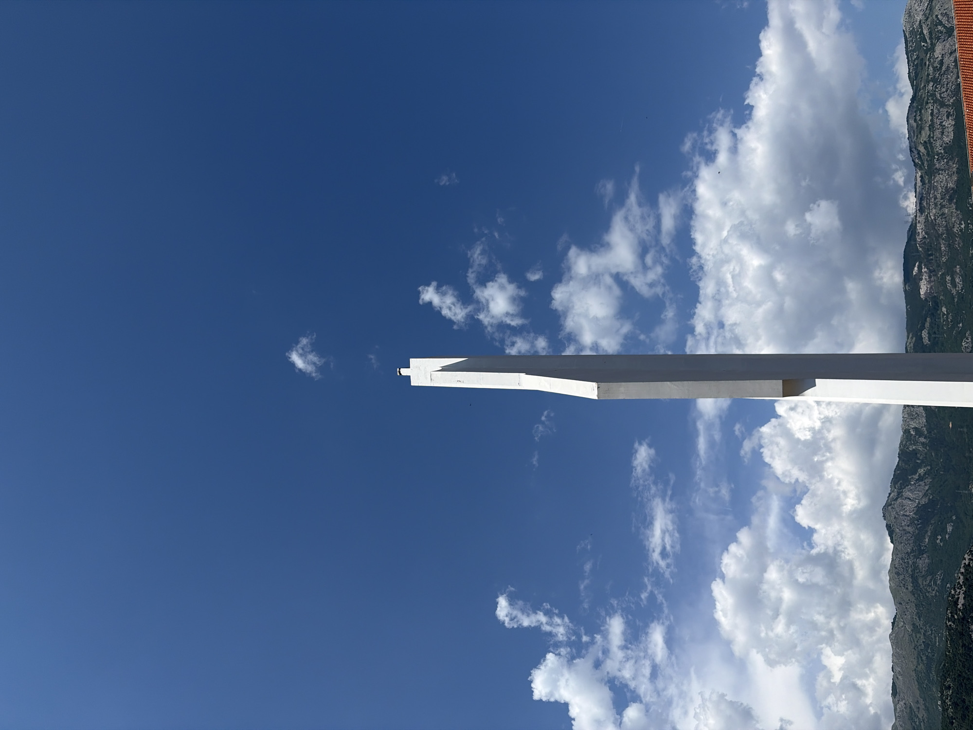 A low-angle shot shows a white concrete structure, possibly a monument or architectural element, against a blue sky with scattered cumulus clouds. The structure appears to be built on a high point, with a mountain ridge visible on the right side of the frame. Some sparse greenery is visible on the side of the mountain. The top of a red-tiled roof can be seen at the edge of the mountain. There are no people or animals directly visible in the scene, although a small bird appears perched on the edge of the top of the concrete structure. This image is likely of the Brajići Cross in Budva, Montenegro.