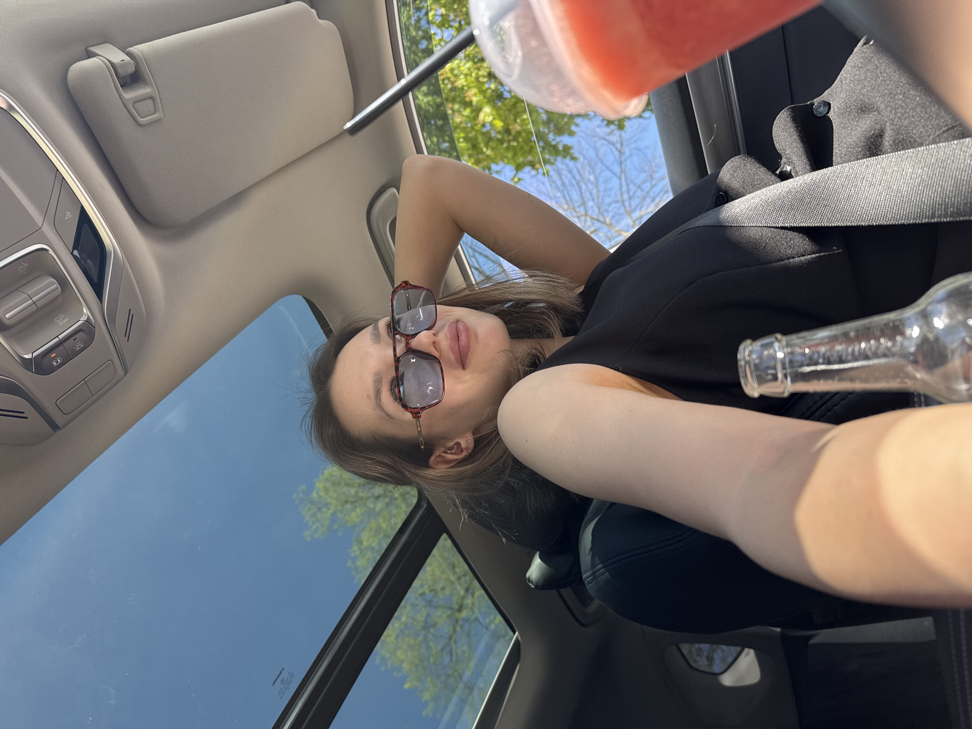 A woman is reclining in the driver's seat of a car, looking at the camera. She is wearing sunglasses and a black top, and is secured with a seatbelt. Her right arm is raised, propping up her head, while her left arm is extended towards the camera, holding an empty glass bottle. To her upper right, a cup containing a red beverage with ice is visible. The car has a sunroof, revealing a blue sky and the tops of trees. The interior of the car features a light-colored sun visor and various dashboard controls. The location is unknown.
