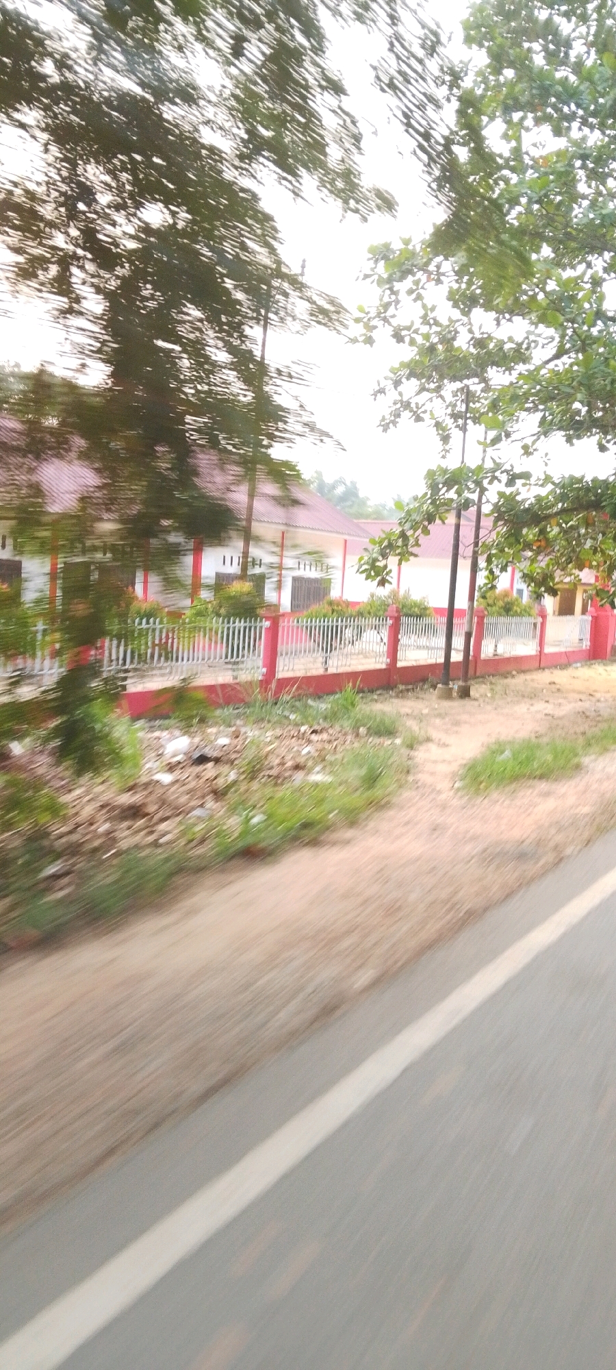 The image, taken from a moving vehicle in Lenggadai Hulu, Indonesia, captures a blurred view of a building with a red-tiled roof and white walls, partially obscured by trees with dark green foliage. A white fence with red posts lines the property. The foreground shows a dirt road with a white line marking its edge. The overall impression is of a sunny day with dappled light filtering through the trees.