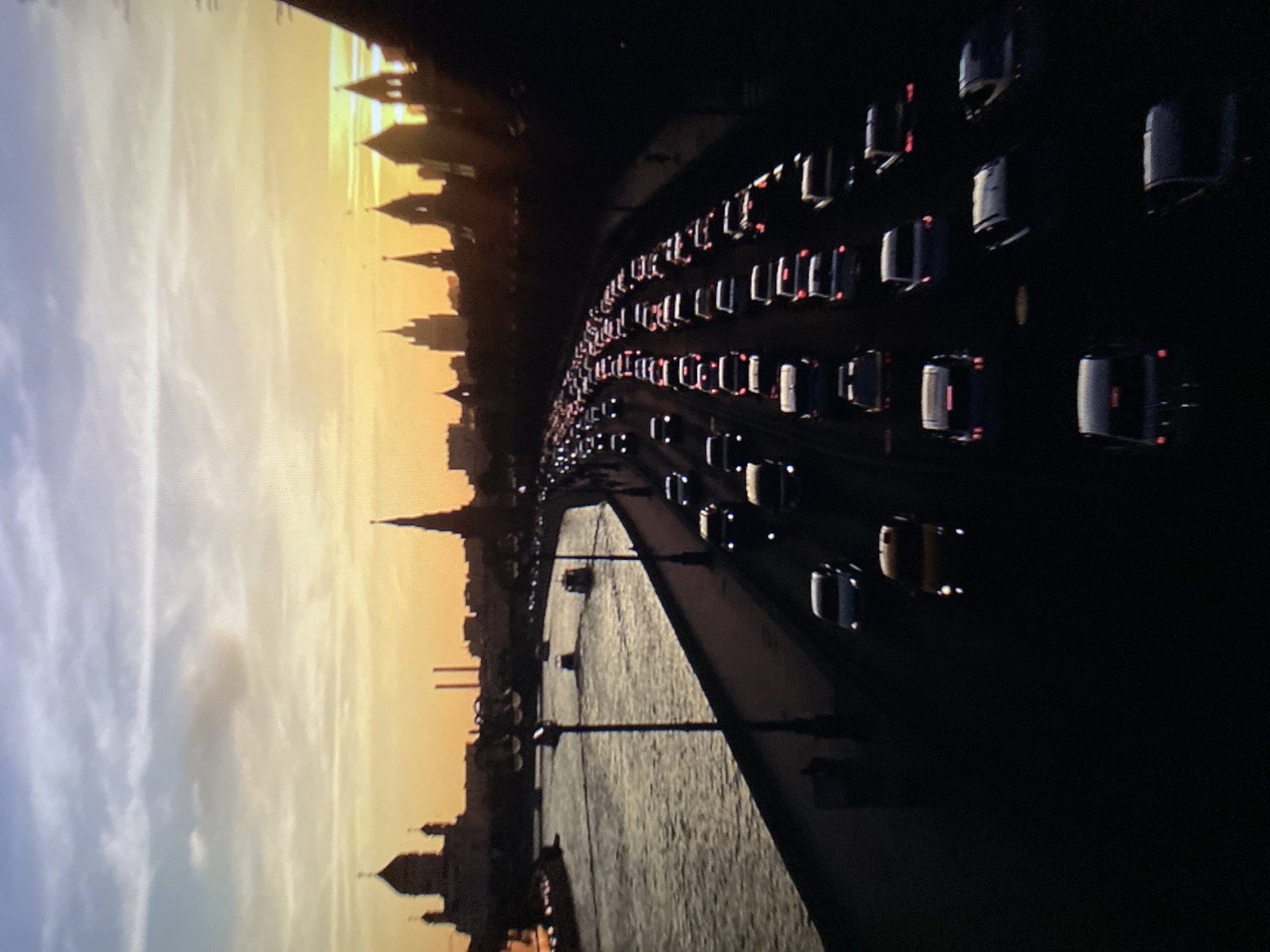 This image depicts a sunset over the city of Khimki, Russia, with a prominent river and a congested highway. The sky is a gradient of blue, purple, and orange hues, with scattered clouds. In the background, the silhouetted skyline of Khimki features several distinct architectural structures, including domes and spires, likely churches or historical buildings.
The foreground is dominated by a wide river on the left, with what appear to be small boats or ferries traversing its surface. A bridge or embankment runs along the river's edge, lined with streetlights.
The right side of the image shows a multi-lane highway heavily burdened with traffic, indicative of a rush hour scenario. Numerous cars are visible, their headlights and taillights creating streaks of light due to the low light conditions. The cars are densely packed, suggesting a traffic jam. The perspective suggests the image was taken from an elevated position, possibly another bridge or a tall building overlooking the highway and river. The overall scene conveys a sense of urban activity and the transition from day to night.