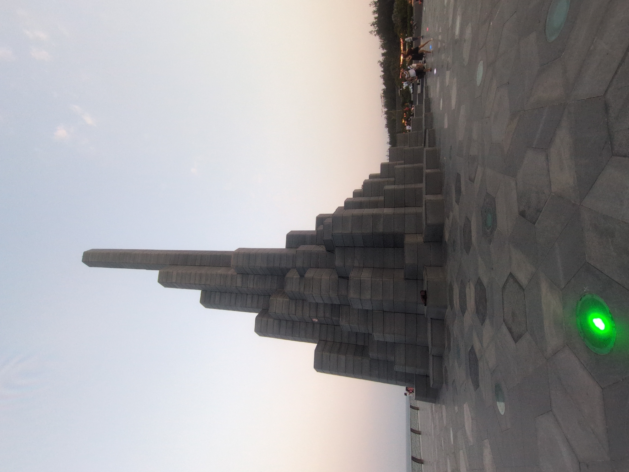 The image shows a large, gray monument composed of stacked, rectangular blocks in Binh Kien, Vietnam. The monument is situated on a paved surface with hexagonal patterns and embedded circular lights, one of which emits a bright green glow. In the background, trees and indistinct figures of people are visible. The sky is a pale, gradient mix of white and light orange, suggesting either sunrise or sunset. The monument's orientation is such that it appears to be pointing towards the sky.
