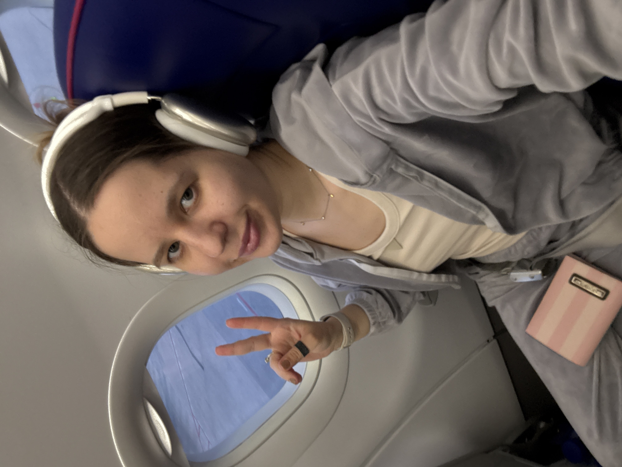 A woman takes a selfie on a commercial aircraft, identifiable by the oval window with a view of a sky, the interior of the cabin and airplane seat. The woman is wearing white headphones, a gray sweatsuit, and a light-colored top. She has dark hair and appears to be making a peace sign with her hand. She has a silver necklace with small charms. In her left pocket, a pink and white striped notebook/planner is visible. The location in the city of El Prat de Llobregat, Spain cannot be precisely identified from the image but given the context of an aircraft could be inferred as being in or around Josep Tarradellas Barcelona–El Prat Airport.