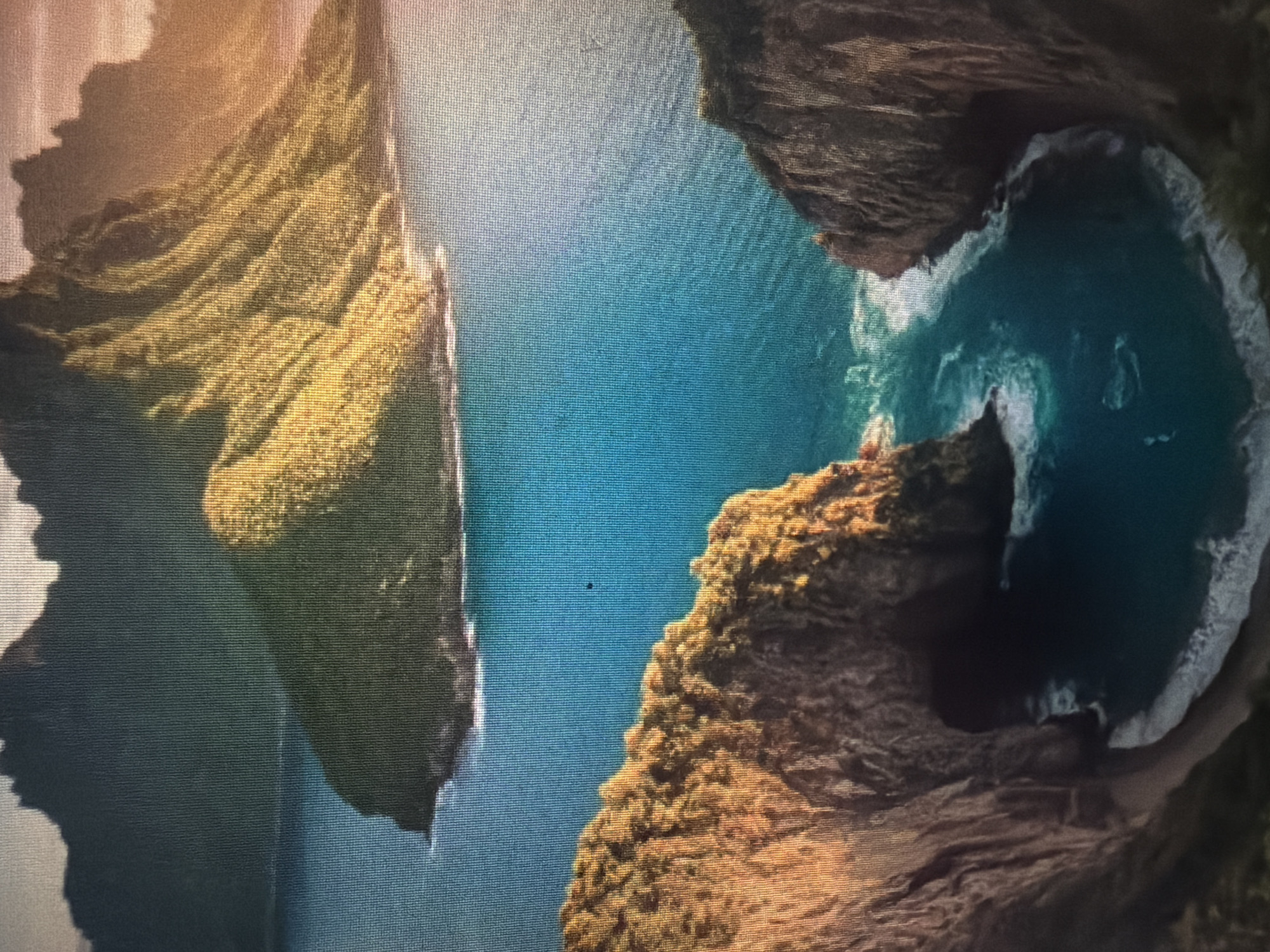 This is a high-angle, full shot of a dramatic coastal landscape, likely a fictional or digitally rendered scene, not a real location in Baan Mae Nam, Thailand. The image features a vast expanse of vibrant blue water, possibly an ocean or a large bay. Dominating the scene are towering, verdant mountains with steep, rugged slopes. The sunlight catches the peaks and ridges of these mountains, casting long shadows and highlighting the textured surfaces.
In the foreground, two imposing cliff faces frame a cove. The cliffs are rocky and uneven, with hints of vegetation clinging to their surfaces. The cove itself contains a small, sandy beach, with gentle waves lapping at the shore. The water within the cove is a deep turquoise, contrasting with the brighter blue of the open water. In the distance, another island or mountain range rises from the sea, its silhouette dark against the hazy, sunlit sky. The overall impression is one of remote, untamed natural beauty.