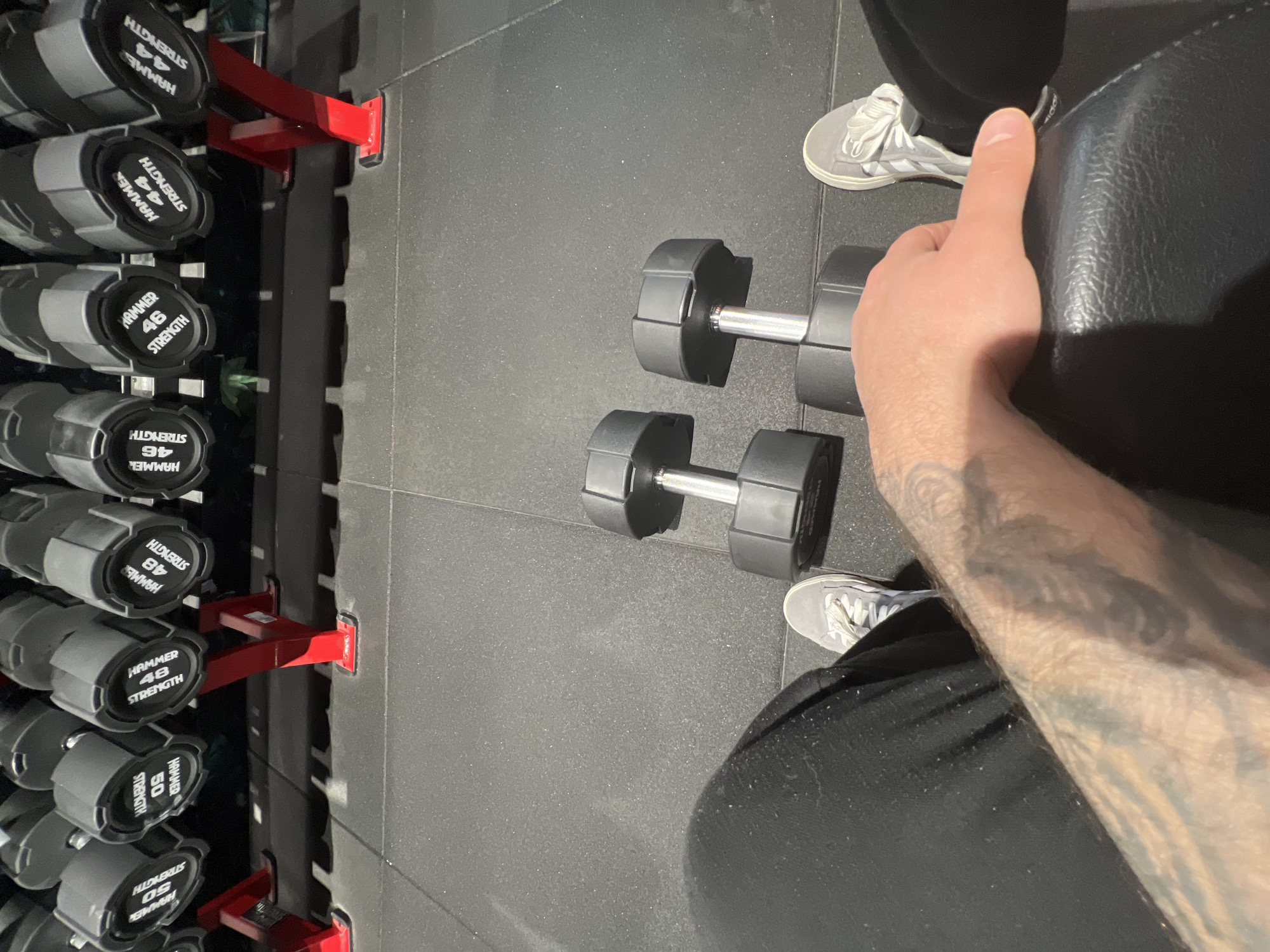 The image presents a view inside what appears to be a gym or fitness center. A dumbbell rack is filled with varying weights of dumbbells, ranging from 44 to 50, labeled with "HAMMER STRENGTH." In the foreground, there are two sets of dumbbells on the floor, positioned near a black weight bench. A person is visible, seated on the bench, with part of their arm and leg visible, showing grey sneakers and black clothing. The person's arm features a tattoo. The floor is dark and appears to be made of a rubber or synthetic material common in gym environments. There is no clear indication of the location being Strasbourg, France, within the frame.