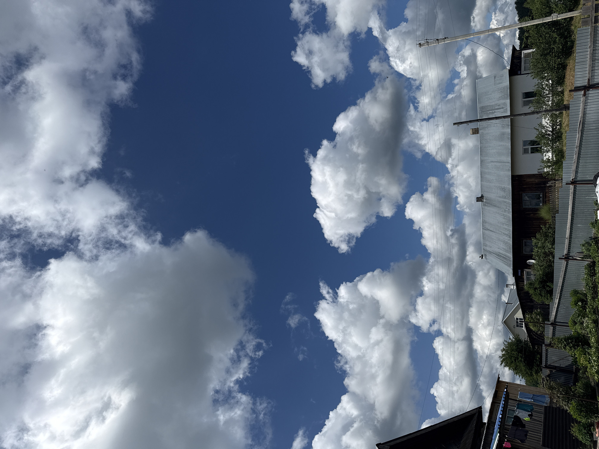 The image shows a daytime scene in Уральское, Russia. A bright blue sky is partially filled with white cumulus clouds. The right side of the frame features rooftops and the sides of buildings constructed of corrugated metal and dark wood, indicative of residential structures. Electrical poles with wires extend into the scene. Foliage including trees and shrubs are also visible amongst the houses. Clothes are hanging on a line in the lower right.