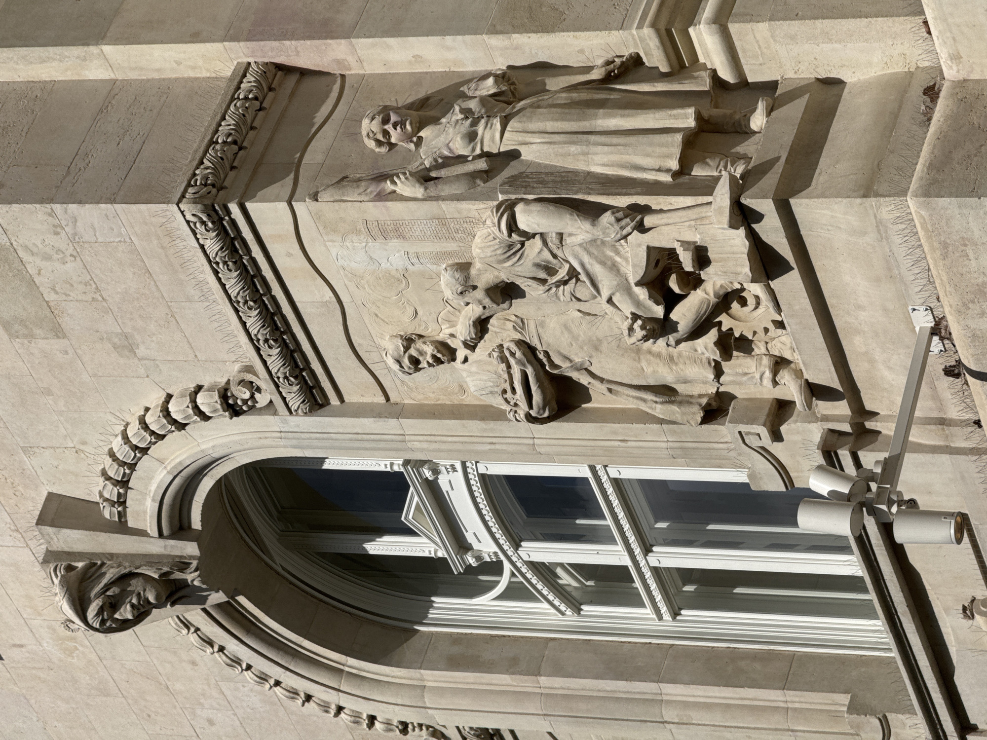 This image captures a detailed section of a grand, ornate stone building facade in Budapest, Hungary, likely viewed from below or an adjacent structure on a sunny day. The sunlight casts strong, well-defined shadows, highlighting the architectural relief work.
The upper portion of the facade features a standing allegorical stone sculpture of a figure, possibly a woman, clad in draped garments and holding what appears to be a scroll or book. Below this, a large stone relief depicts multiple figures engaged in an activity that suggests labor or craftsmanship. One prominent figure is seen crouching or kneeling, with another figure's head partially visible in the lower left of the relief.
The building exhibits elaborate stone carvings, including decorative friezes with intricate foliate motifs. A large, arched window with white frames, possibly multi-paned, is visible in the lower left, appearing slightly ajar. Above this window, a grotesque head sculpture protrudes from the ornate archway.
Contrasting with the historical architecture, a modern security camera or lighting fixture, consisting of several cylindrical units on a pole, is attached to the building on the right side of the window. Anti-bird spikes are also visible along some of the ledges and cornices, indicating maintenance efforts to preserve the building's detailed stonework. No legible text is visible.