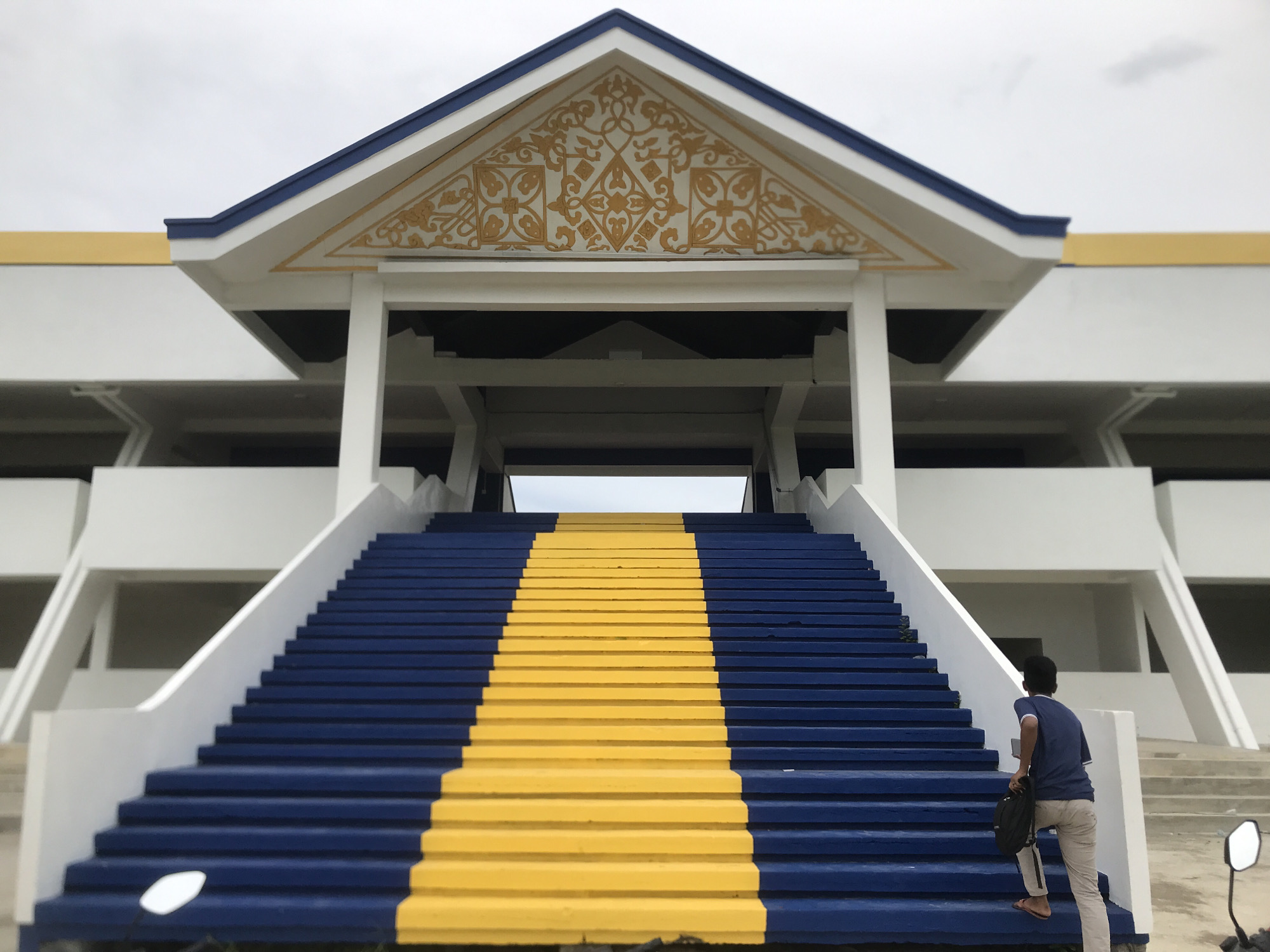 A medium-angle shot depicts a man ascending a large staircase in Tambontunong, Indonesia. The staircase, painted with alternating blue and yellow steps, leads to a building with a distinctive triangular pediment adorned with gold filigree. The building is predominantly white with blue trim. The man, dressed in a blue shirt and tan pants, carries a dark bag and is partially visible from the side. A motorcycle mirror is visible on the lower right. The sky is overcast.