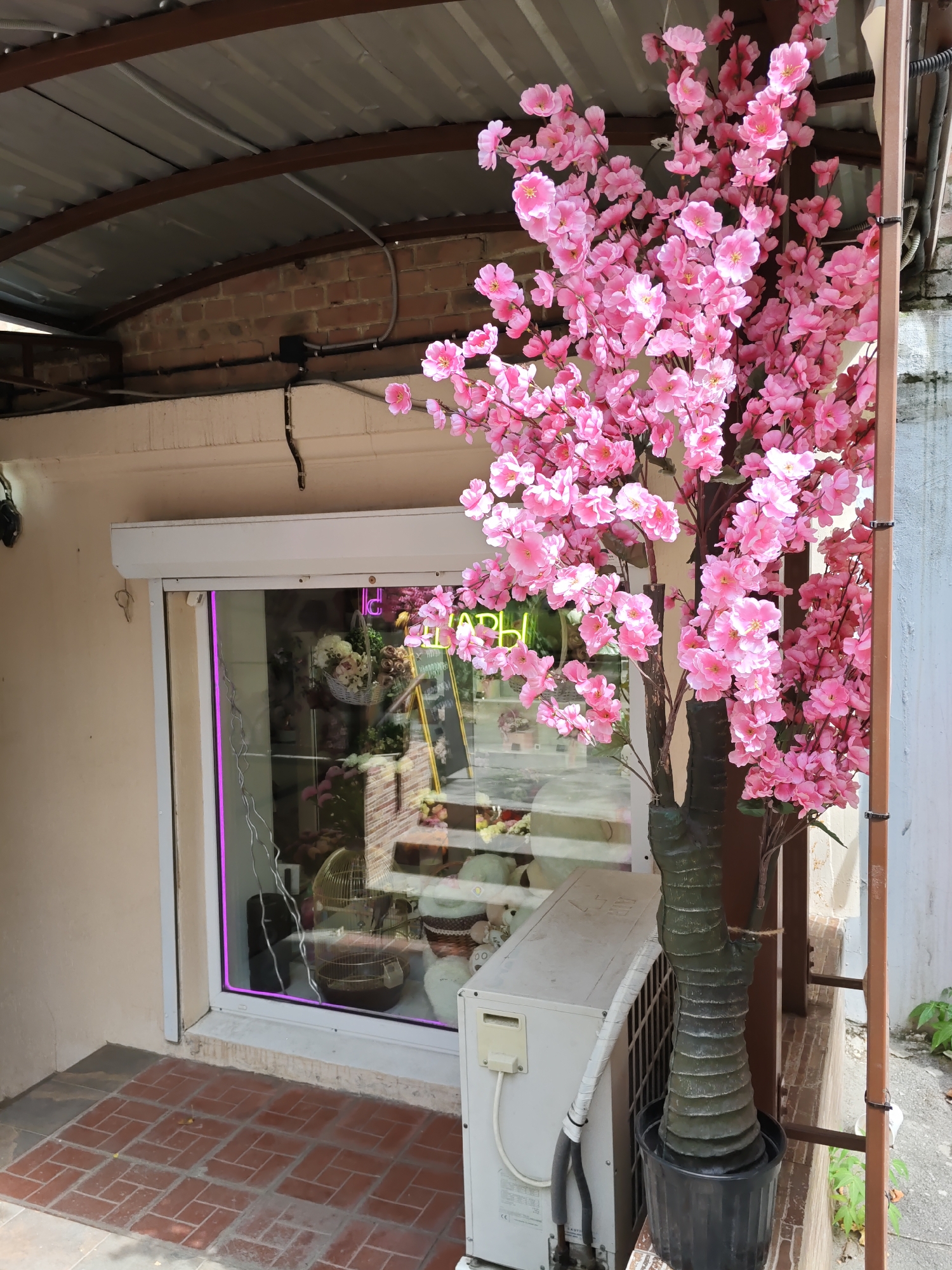 In Rostov-on-Don, Russia, a storefront is depicted with a prominent artificial cherry blossom tree in the foreground. The tree, with its vibrant pink flowers, stands in a black pot. To the left, a shop window displays various items, including floral arrangements, decorative birds in cages, and teddy bears, with neon pink lighting framing the window. An air conditioning unit is situated below the window. The storefront itself has a light-colored facade, and the ground in front is paved with red brick. Above the storefront, a corrugated metal roof is supported by metal beams, with exposed brickwork visible beneath. Electrical conduits and wiring are visible along the wall and under the roof. A metal ladder or support structure is partially visible to the right of the cherry blossom tree.