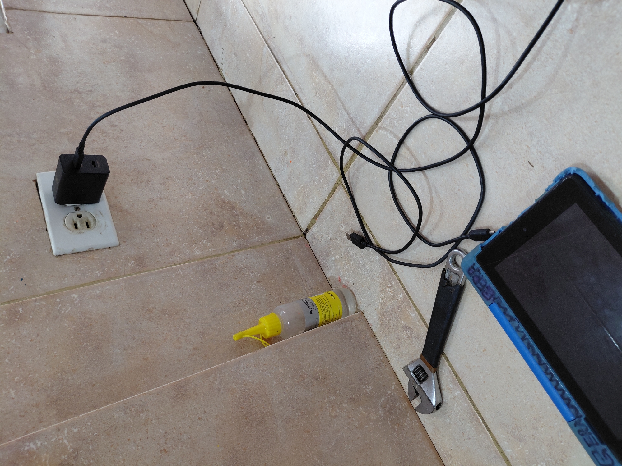 This image depicts an indoor scene in Santa Ana, Venezuela, featuring electronic devices and tools resting on a light brown, tiled floor. A black power adapter is plugged into a white electrical outlet on a wall. A black USB cable extends from the adapter, winding across the tiled surface towards a tablet, suggesting it is being charged. The tablet is housed in a worn blue protective case, on which the text "GERA" is faintly visible and repeating along its edge.
Adjacent to the tablet lies an adjustable wrench with a silver head and a black handle. Further along the tiled floor, near a corner where two tiled sections meet, is a clear plastic bottle with a yellow cap and nozzle. This bottle contains a white substance, and its yellow label is clearly visible, displaying the text "SILICONA LÍQUIDA" (Liquid Silicone), "SELLADOR MULTIUSOS" (Multipurpose Sealant), and "CONT." (Content).
The tiled floor shows signs of wear, with visible grout lines and some minor dirt or stains. The white electrical outlet cover also appears somewhat discolored and used. The overall arrangement suggests a casual, everyday environment, possibly a workspace or area for charging devices and incidental tasks.