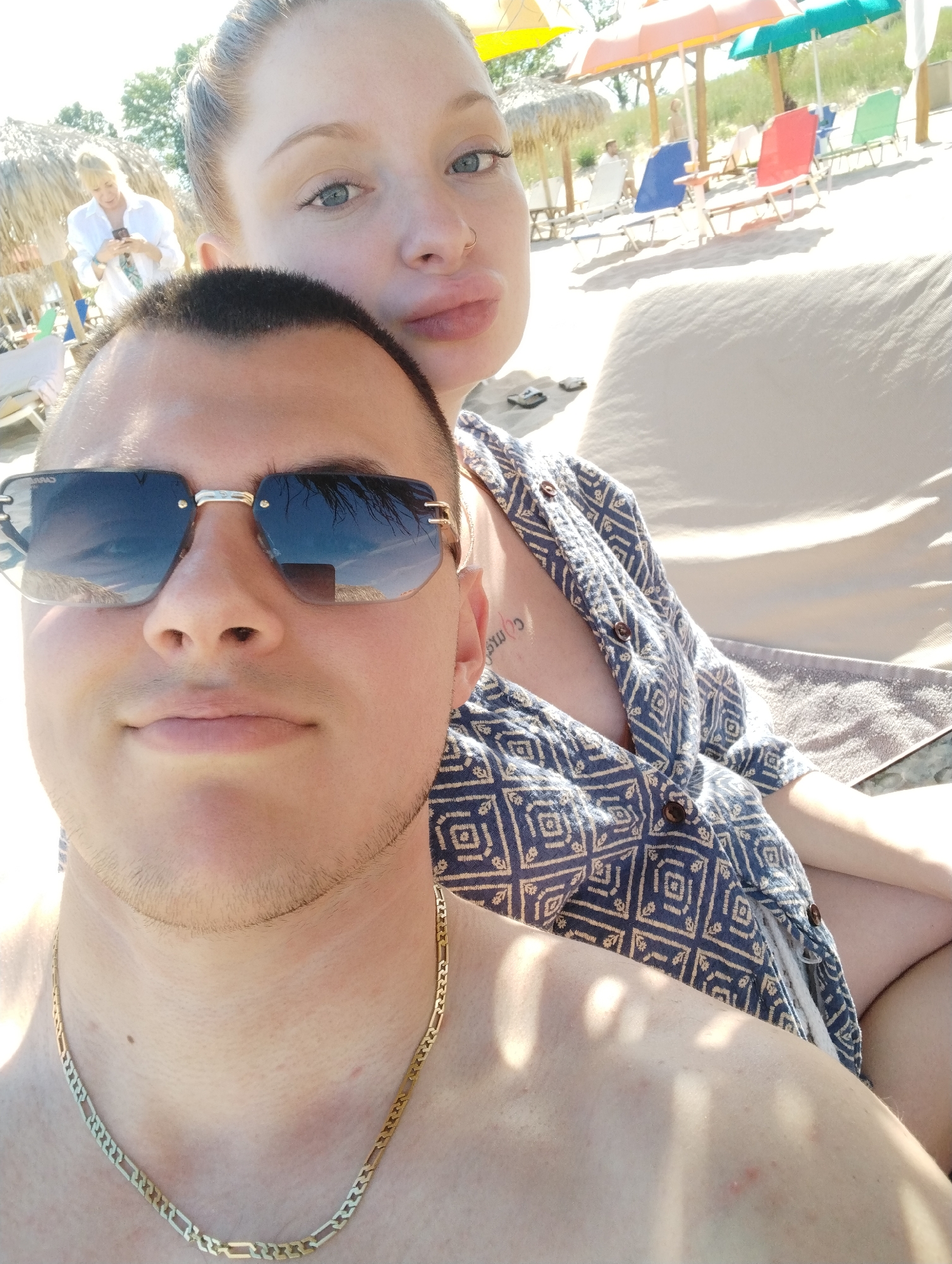 The image shows a selfie of a man and a woman on a beach. The man is in the foreground, wearing sunglasses and a gold chain. The woman is behind him, with her head tilted back and her lips pursed. She is wearing a blue and white patterned shirt. In the background, there are beach chairs, umbrellas, and a few other people. The scene appears to be a sunny day at the beach.