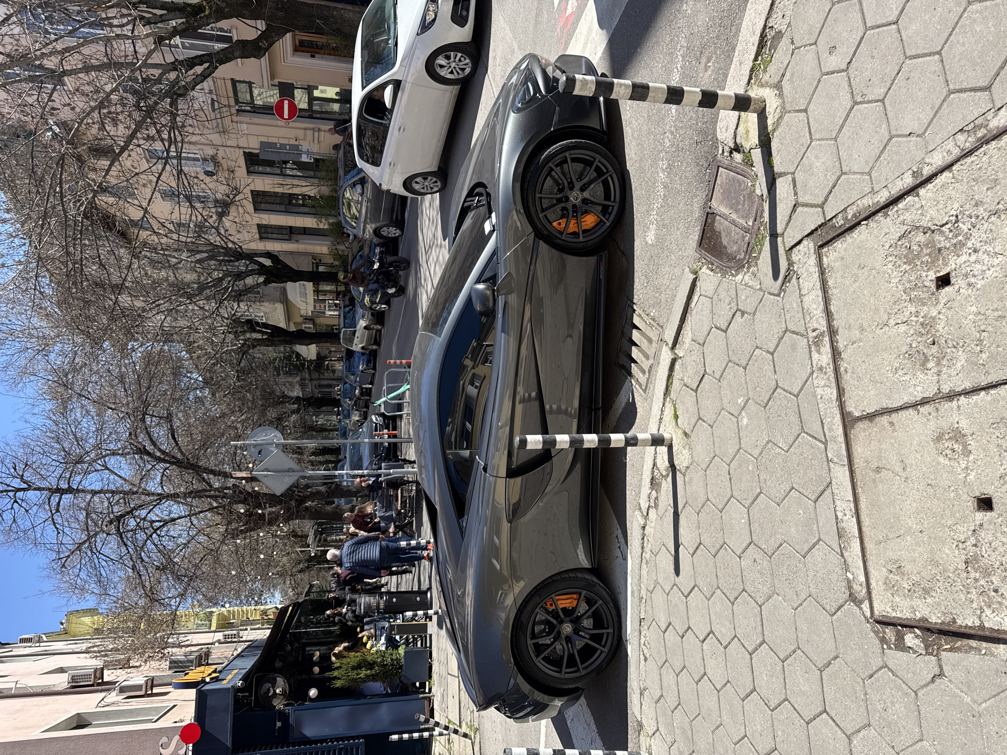 The image captures a street scene in Sofia, Bulgaria. A gray McLaren sports car is parked on the right side of the street. Black and white striped bollards stand nearby. Other parked vehicles, including a white van, can be seen in the background. Pedestrians are walking along the sidewalk, passing by an outdoor cafe with blue awnings and red accents. Bare trees line the street, and buildings with beige facades are visible. A street sign is also present. The pavement is made of hexagonal blocks.