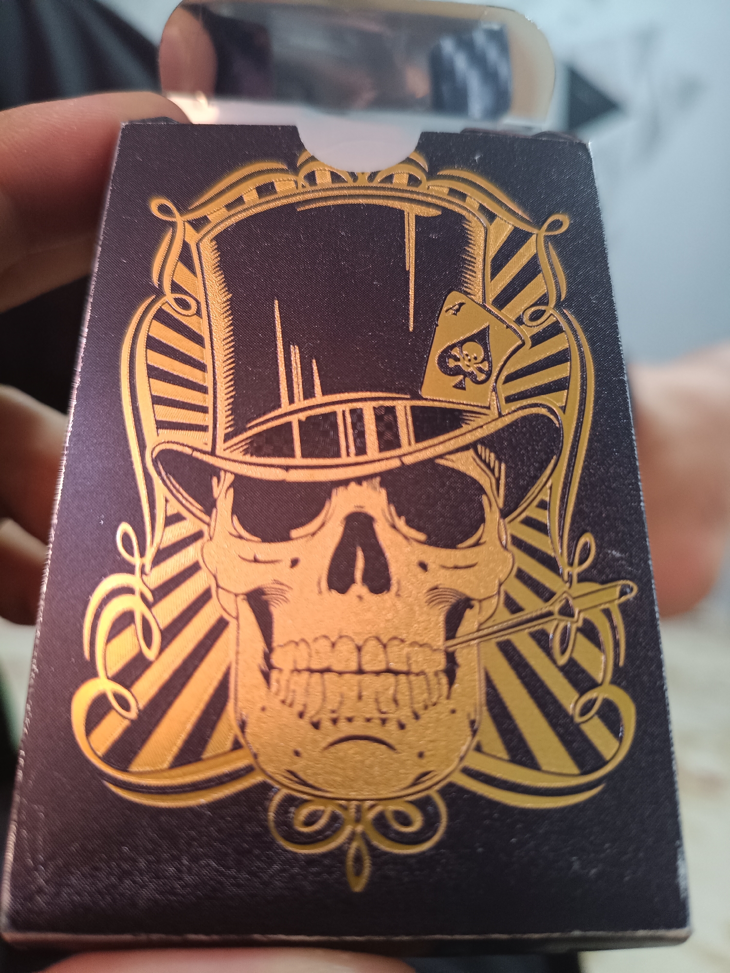 A close-up, slightly angled shot shows a person's fingers holding the front of a dark blue playing card box with a gold embossed design. The design features a skull wearing a top hat, with a cigarette in its mouth. An ace of spades card with a skull emblem is positioned on the brim of the hat. The background is blurred, showing what appears to be a person's hand or arm in the upper left and right corners, and a light-colored surface in the background. The card box has a textured surface and appears to be made of thick cardboard or paper. There is no discernible information about the location in Aktau, Kazakhstan.