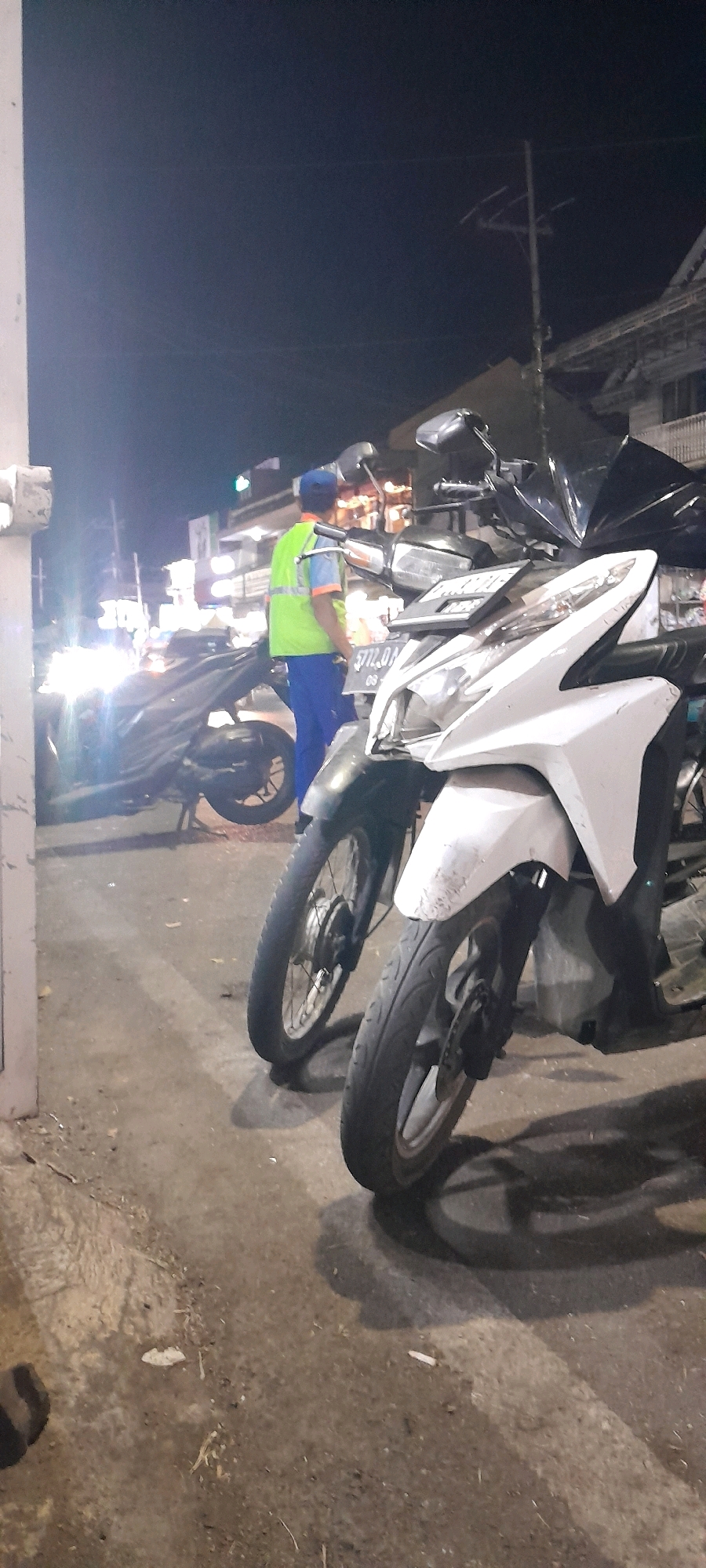 In Rogojampi, Indonesia, at night, a person wearing a reflective vest and blue pants stands near a parked motorcycle. The motorcycle in the foreground is white, with its front wheel visible and a license plate that reads "5772 OM 08". Behind it, another motorcycle is parked, appearing darker. The background shows a street scene with blurred lights, possibly from shops or streetlights. The overall environment suggests a public area in a town or city at night.
