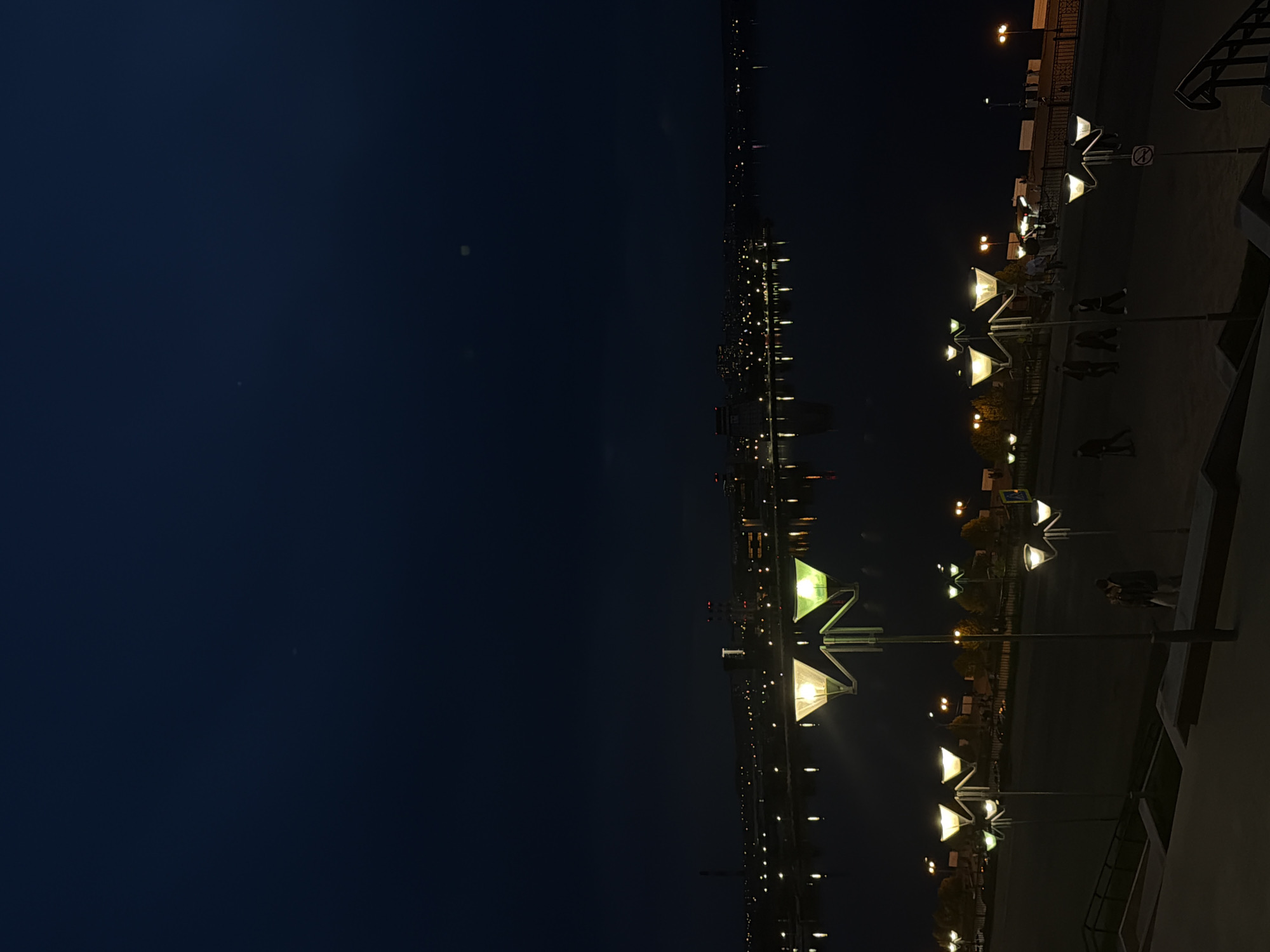 The image depicts a nighttime scene in Izhevsk, Russia. The right side of the image features a promenade or walkway illuminated by a series of distinctive streetlights with a unique geometric design. Pedestrians are visible walking along the walkway. On the left side is a dark expanse, possibly representing the night sky or a body of water, and in the distance, the city lights are reflected on the water. The overall atmosphere is dark, with the streetlights and distant city lights providing the only illumination.