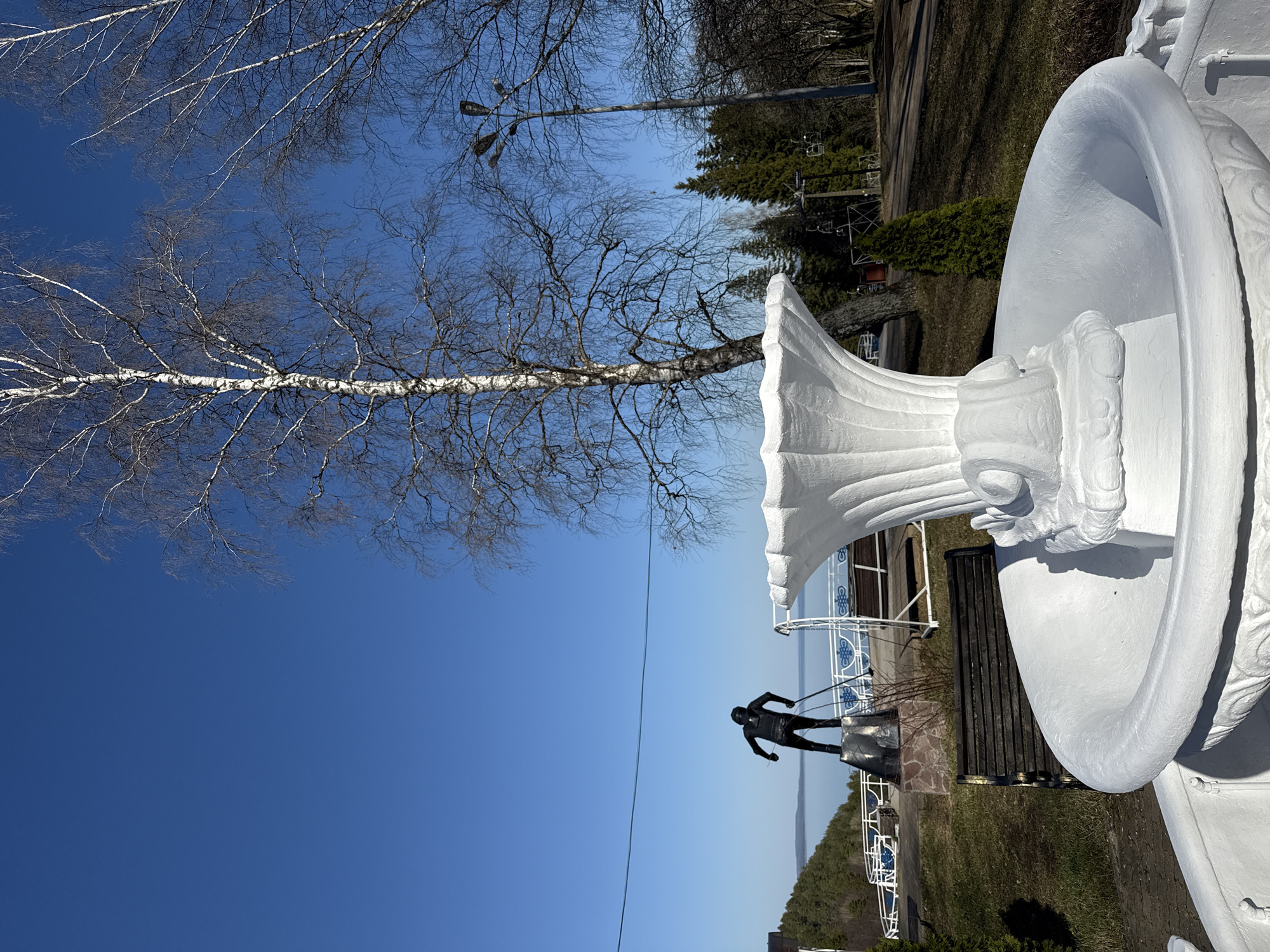 A low-angle view captures a stark contrast of urban elements against a natural backdrop, potentially in Galevo, Russia. A white, ornate vase-like structure dominates the foreground, positioned to the right. To the left, a skeletal birch tree reaches towards a cloudless blue sky. In the middle ground, a dark sculpture of a figure pulling something, possibly a skier, stands on a stone pedestal. A metal railing separates the figure from a body of water with a distant shoreline. Beyond the sculpture, a bench sits adjacent to the vase, with a backdrop of trees and a lamppost. The scene suggests a public park or monument area.