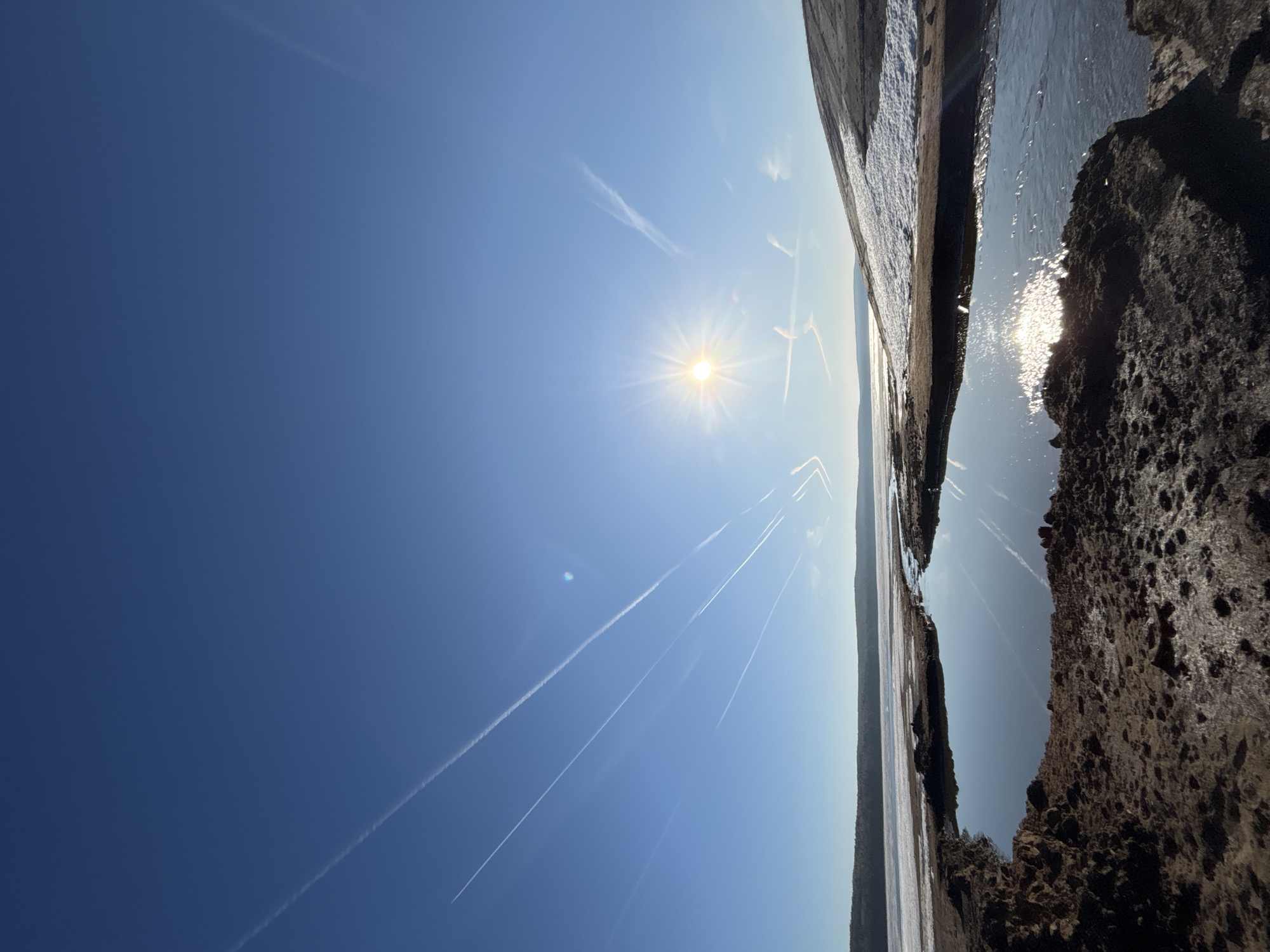 The image shows a daytime view of the sky over a body of water. The sky is clear blue with visible contrails from aircraft. The sun is prominent in the sky. The water surface reflects sunlight and the contrails. The foreground shows a rocky shoreline. The photo was taken near the city of Chaikovsky, Russia.