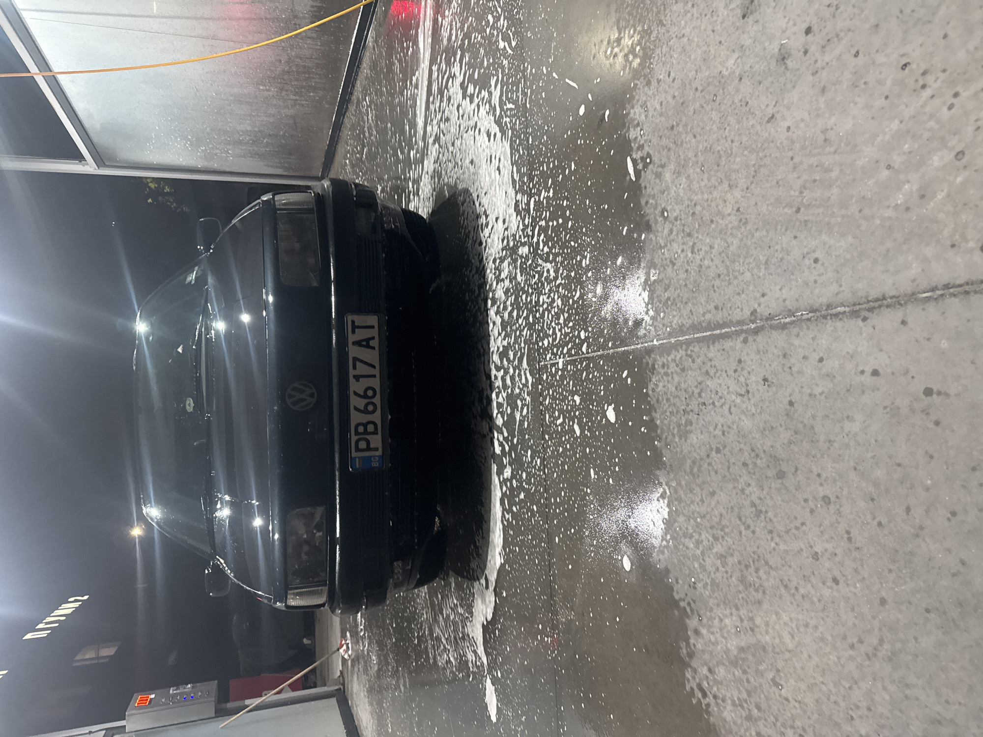 The image shows a black Volkswagen car being washed at a car wash facility in Plovdiv, Bulgaria. The license plate visible on the car reads "PB 6617 AT". The car is covered in soapy water. The car wash appears to be mostly enclosed with some open areas. The ground is wet and covered with soap suds. The lighting suggests it is evening or night. A sign with Cyrillic script is visible in the background. A payment kiosk with a digital display is also present.
